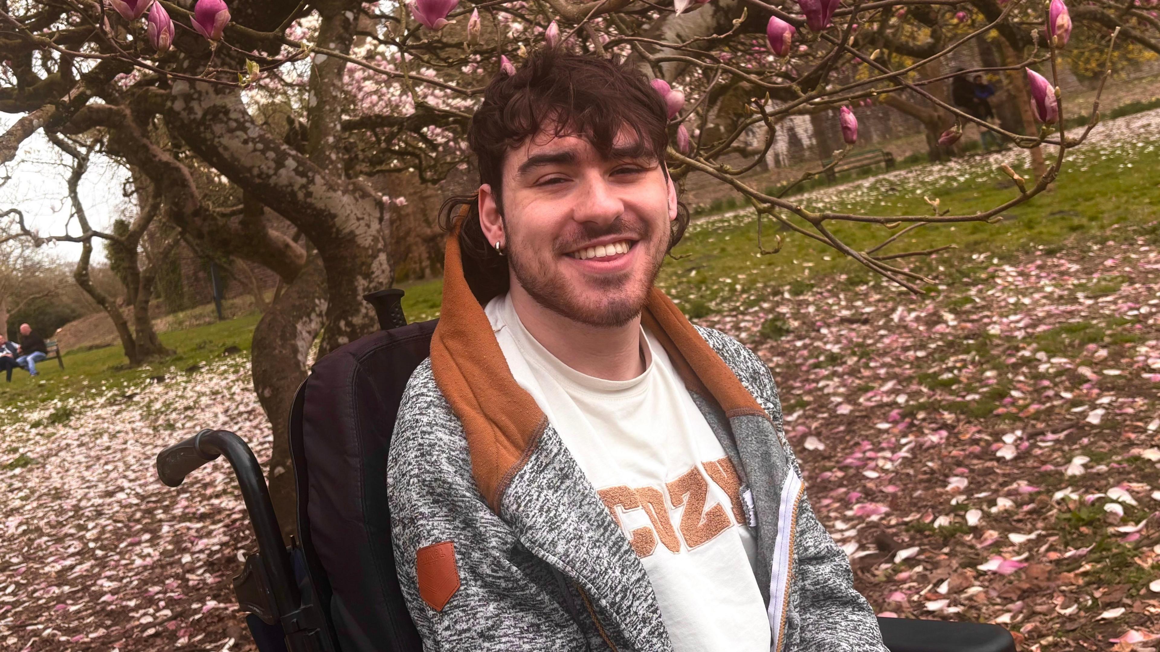 A smiling young man is seated outdoors in a park beneath blooming trees with pink flowers and fallen petals covering the ground. He has dark wavy hair and a light beard, and is wearing a gray and brown hoodie over a white shirt that says cozy, the backrest of his wheelchair is visible behind him.