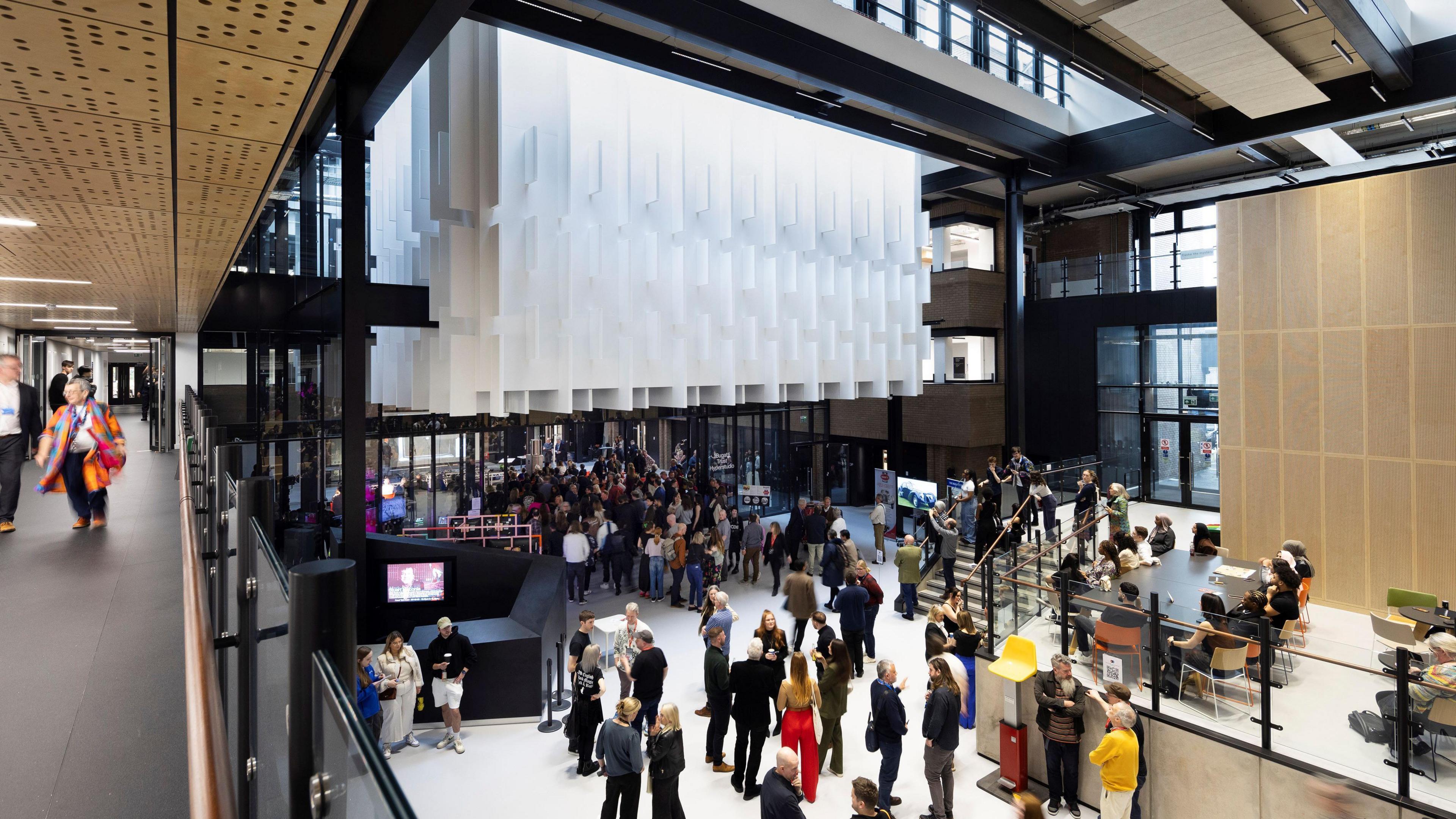 The ground floor of a modern university building pictured from a first-floor balcony. The ground floor has a white jigsaw-like structure above the entrance and lots of people are gathered.