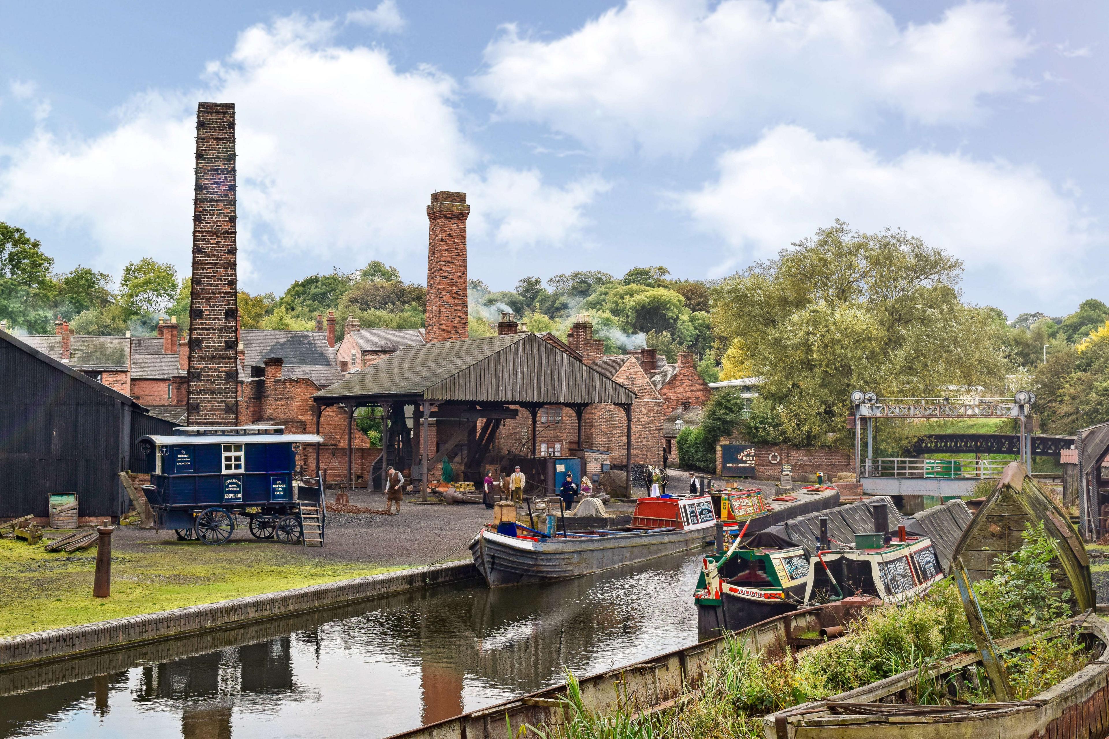 A picture of the canal at the Black Country Living Museum. Three boats are in the water. The right-hand side of the water is covered with green plants, while across the water people are working. There are red-brick buildings and large wooden shelter.