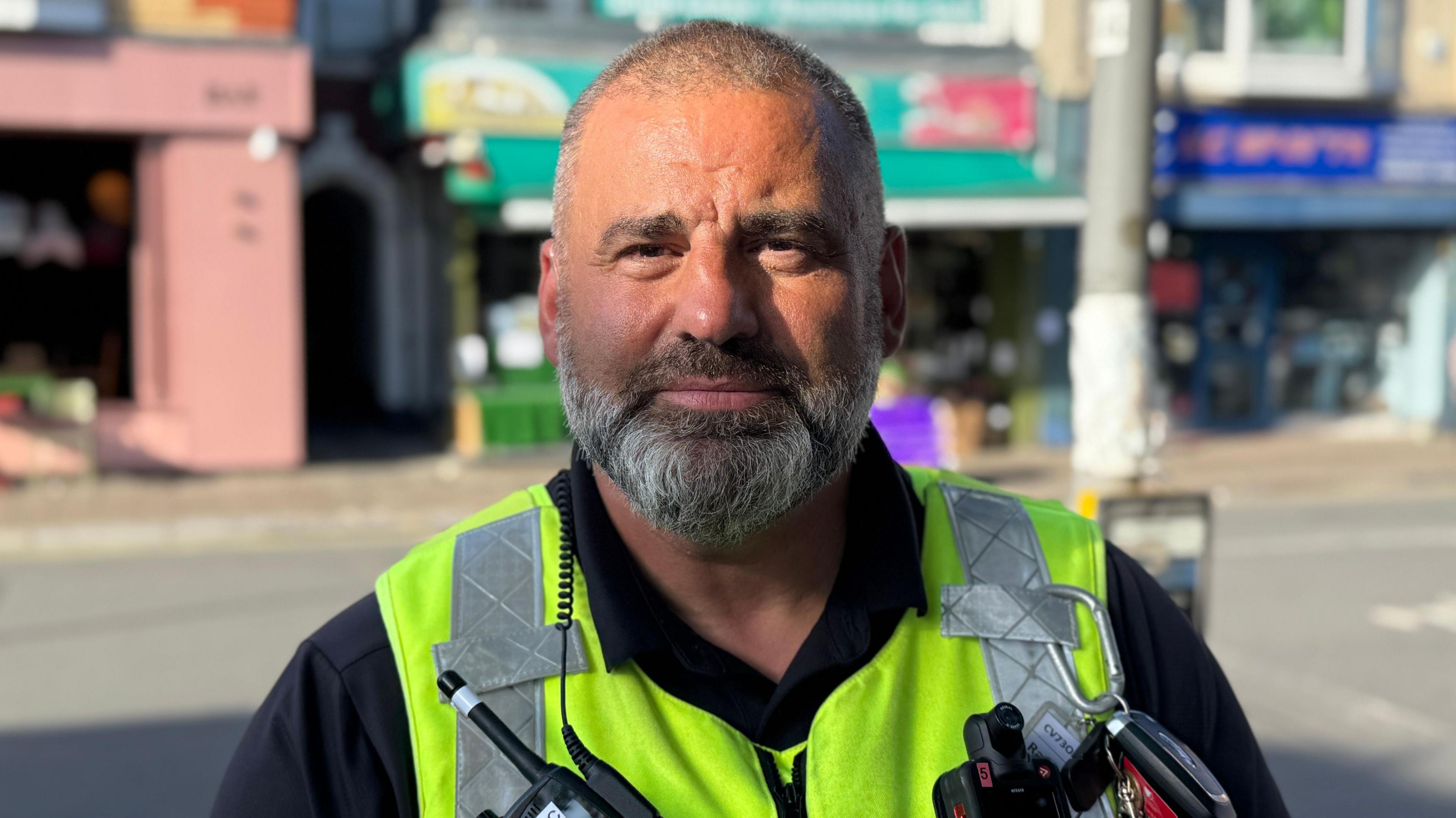 Dave, a man with a shaven head and grey beard wearing a black shirt and a yellow hi-vis vest looks directly into the camera. He is also wearing a radio with an earpiece and a body worn camera. He is standing near a sunny high street.