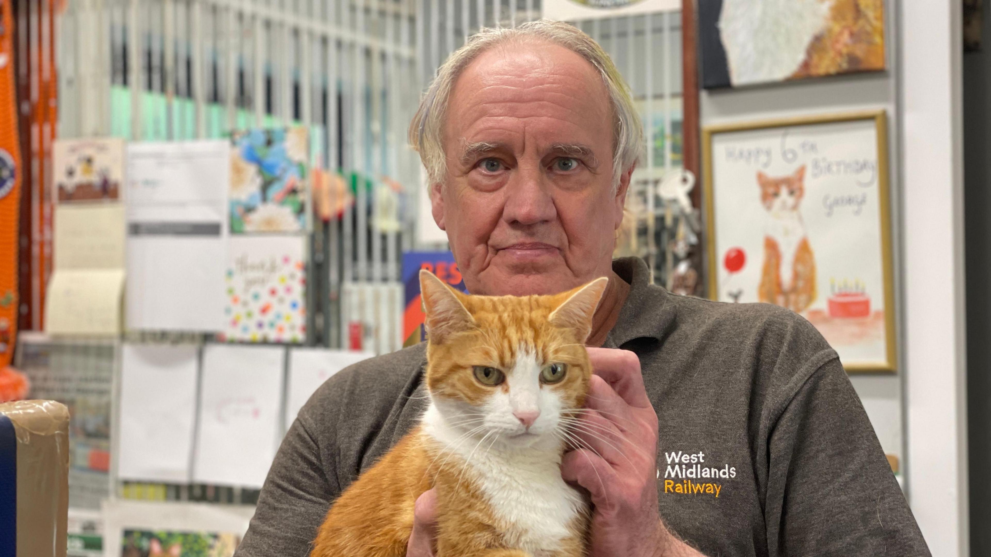 A ginger and white cat being held by a man in a grey polo shirt that has West Midlands Railway embroidered on the left side. Behind there are pictures and calendars visible on the wall, including a drawing of the same cat