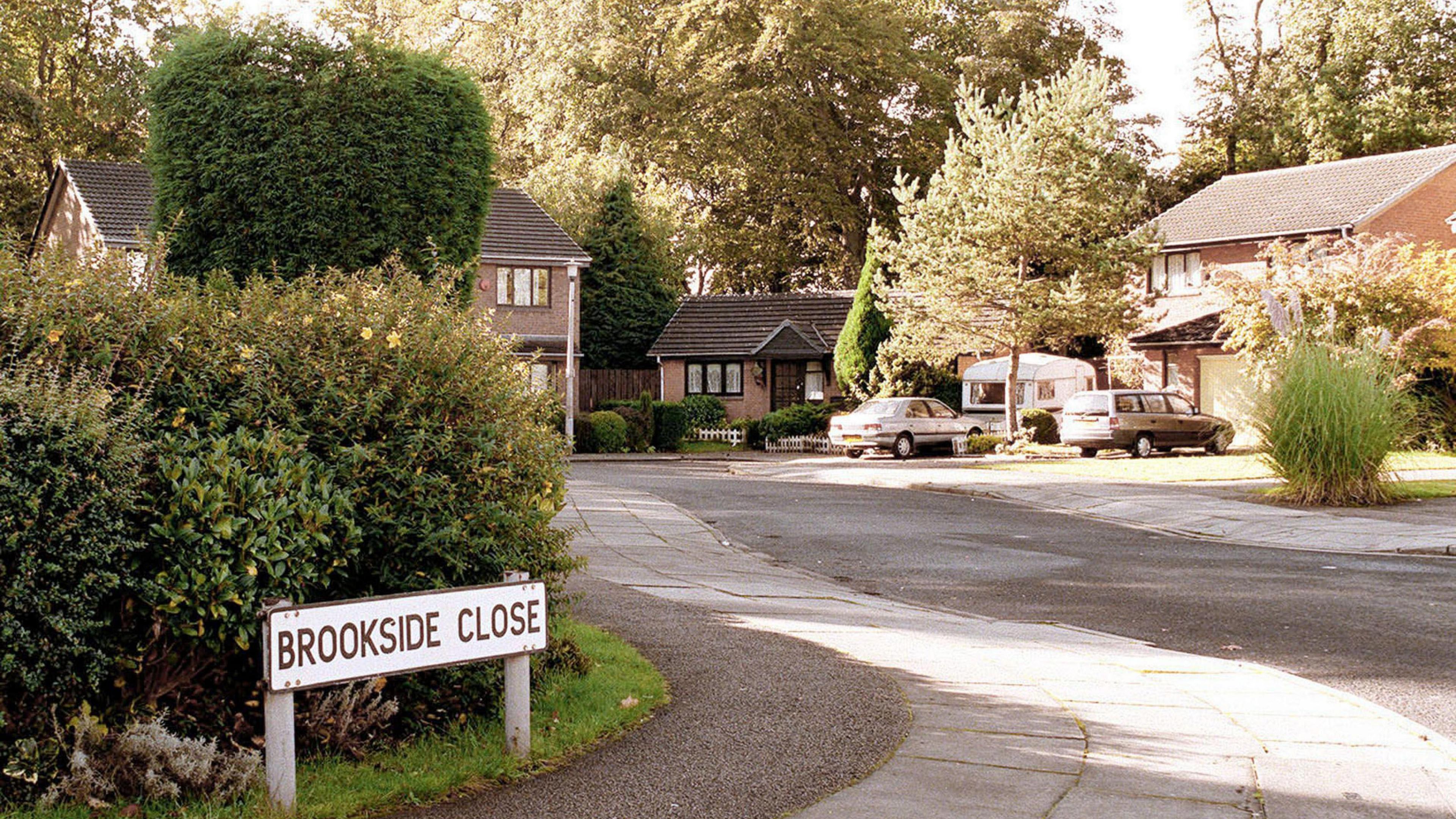A picture of Brookside Close, captured in the 1990s. To the left is a street sign which reads: Brookside Close with bushes behind it. The road curves round to the left with a number of houses and one bungalow in view