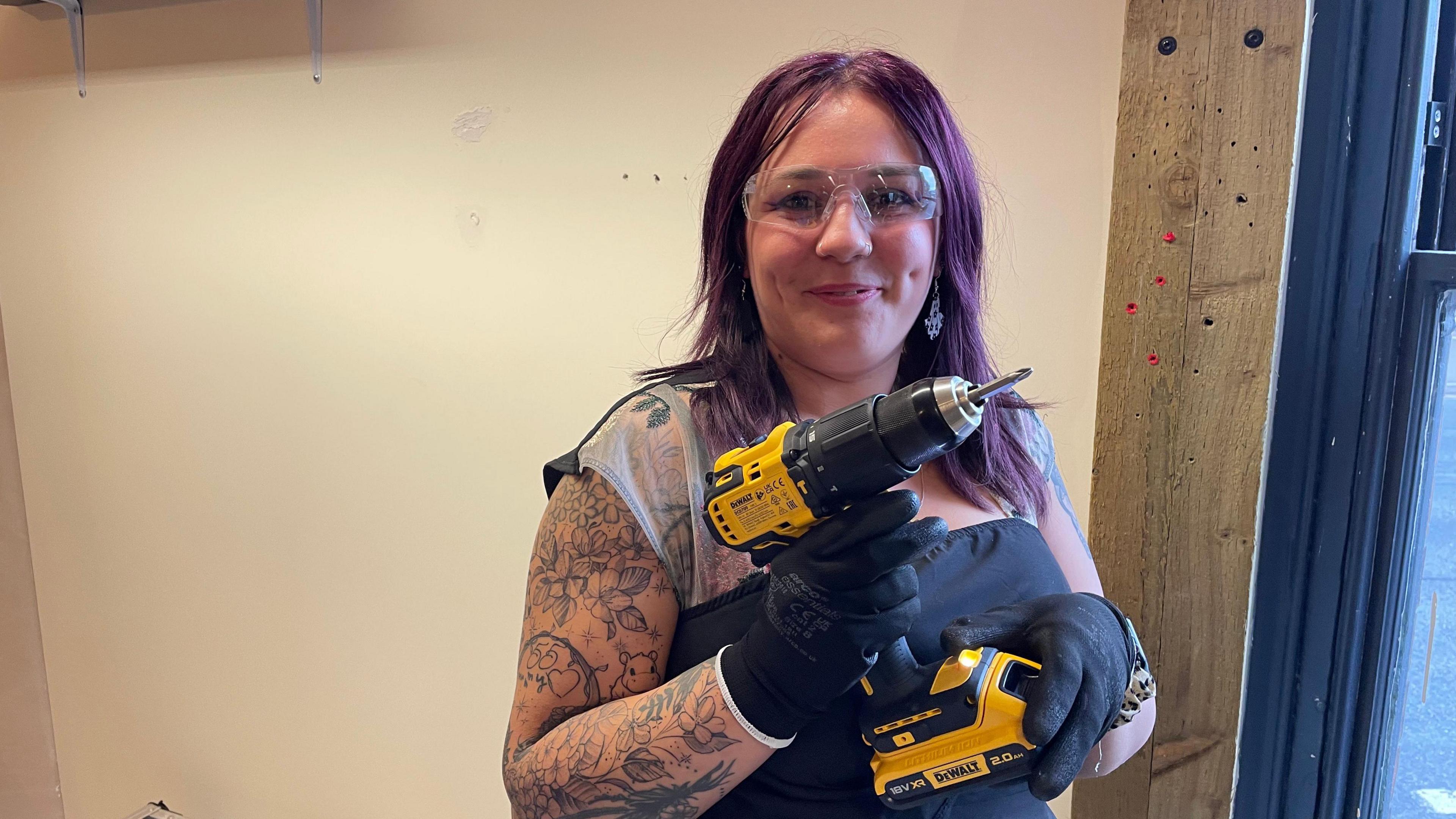 Hope has long purple hair and is holding an electric drill above her arm that is covered in tattoos