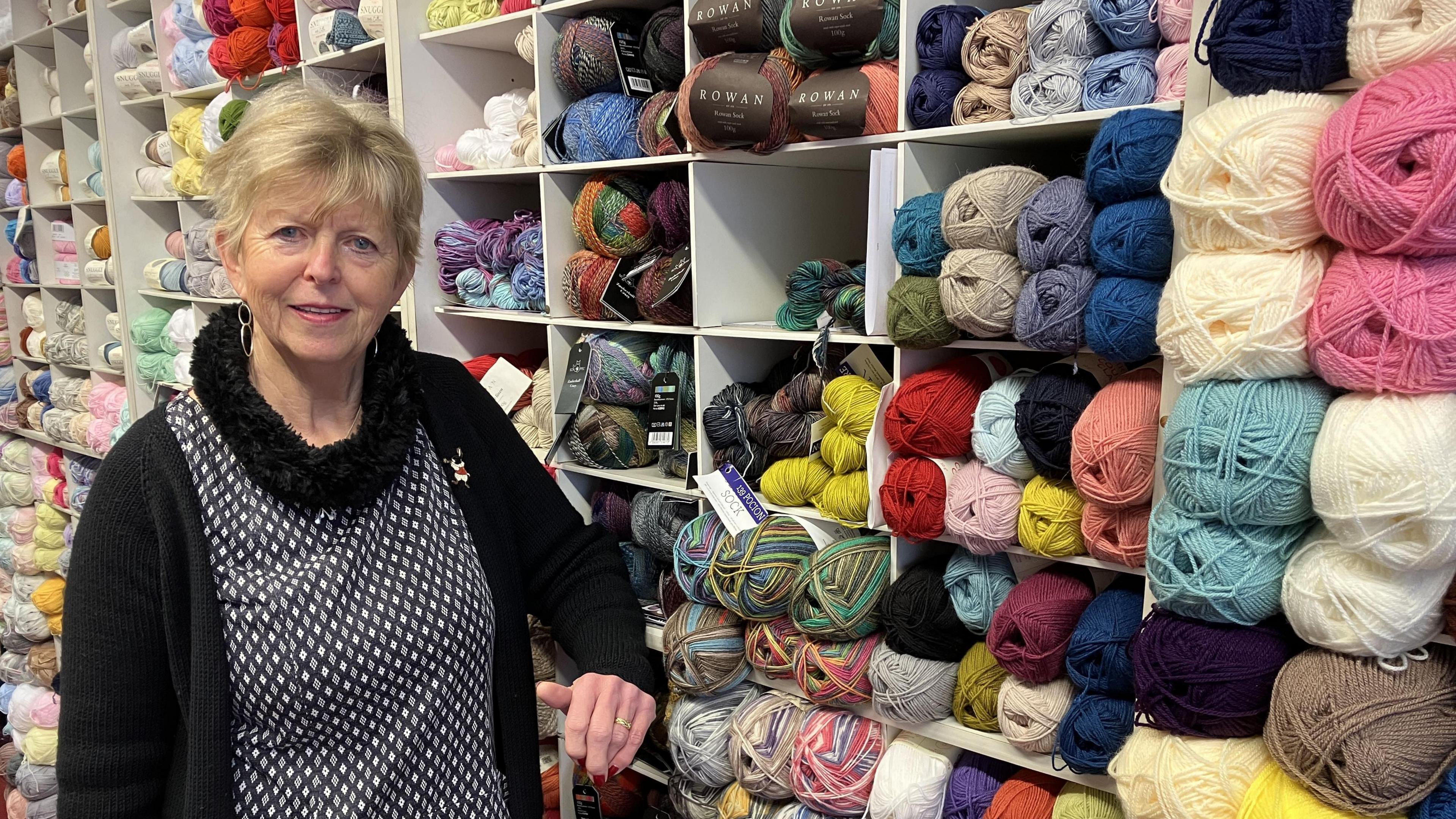 Cynthia Binks who has short blonde hair and is wearing a black and white top, stood in front of shelves stacked full of wool in a huge variety of different colours