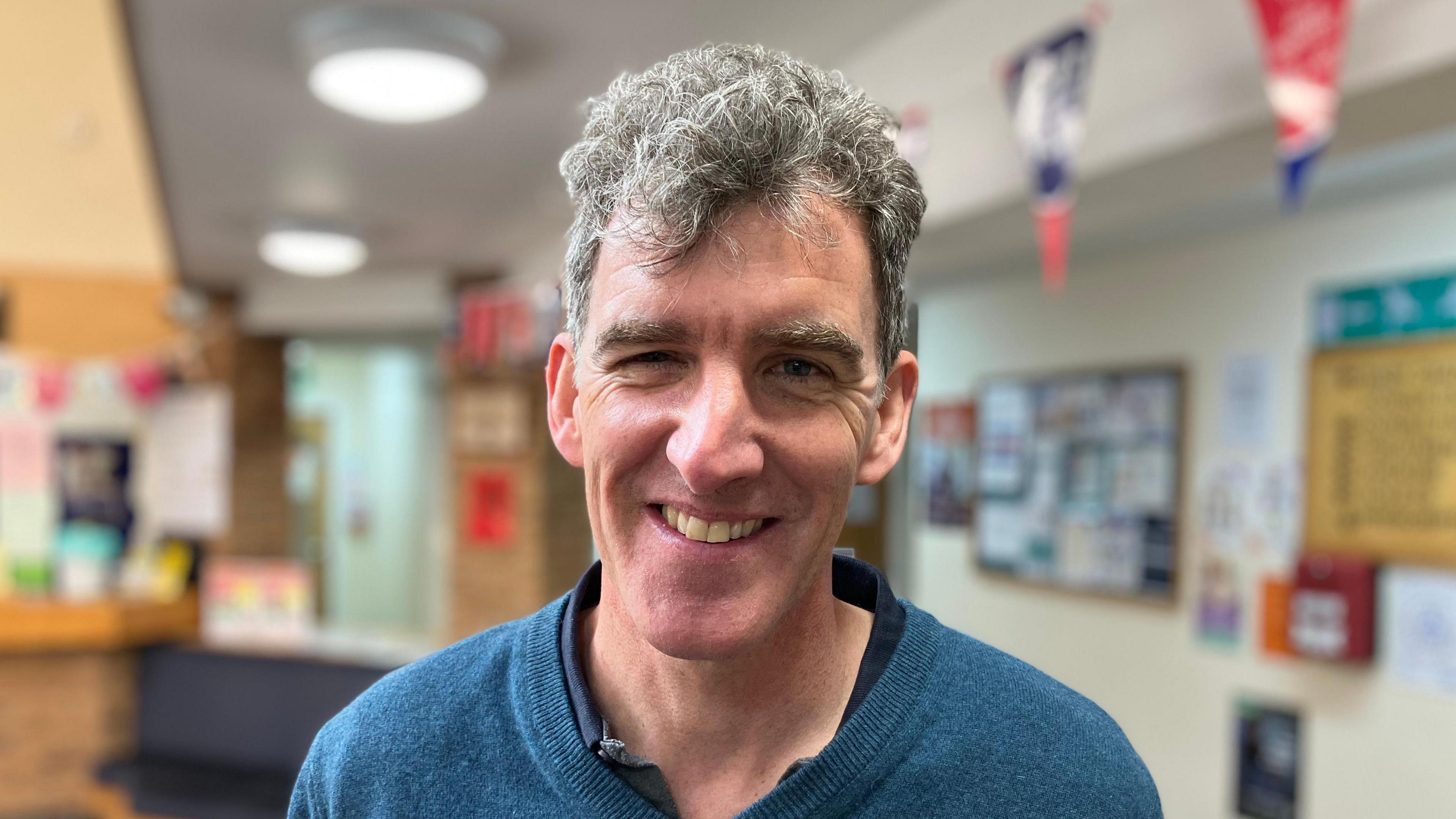 A smiling man, with short, grey hair, wearing a blue pullover over a darker blue shirt