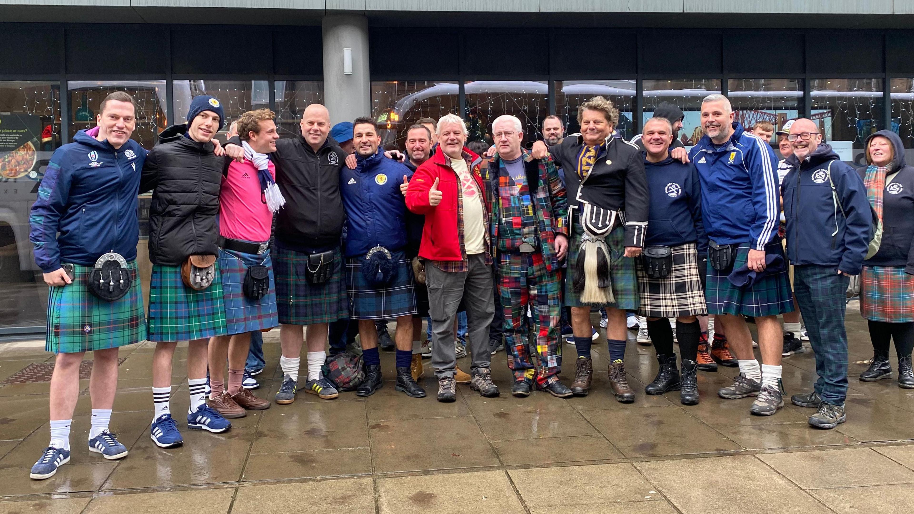 The image shows a group of people standing together outdoors on a wet pavement in front of a modern building with large glass windows. The group are wearing  a mix of traditional Scottish attire and casual clothing.