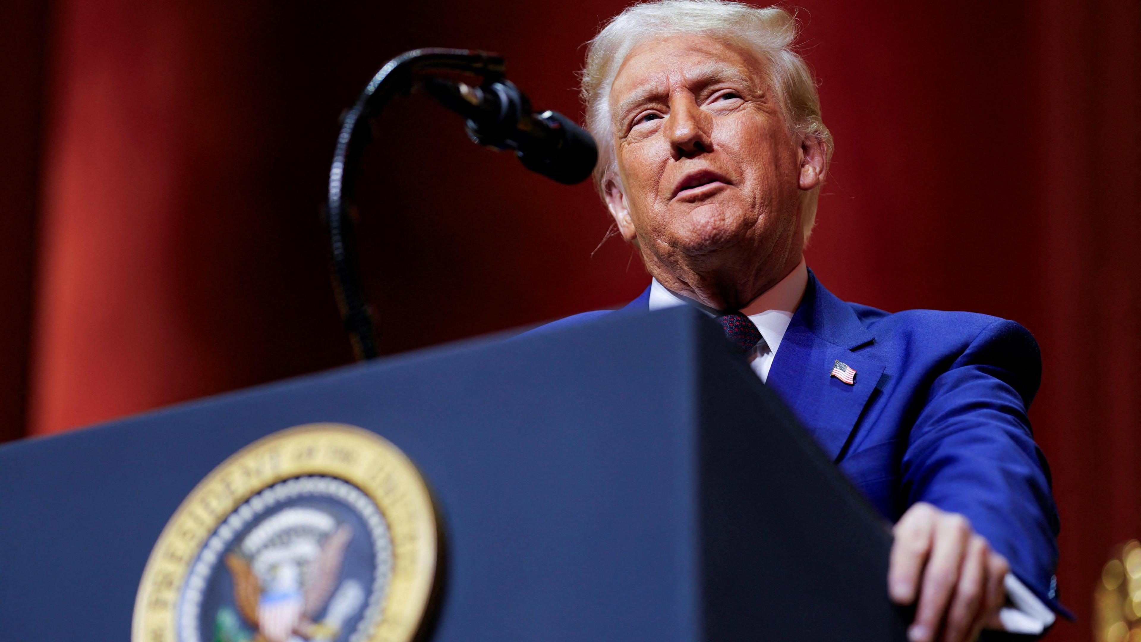 Donald Trump speaks at a podium. He has grey hair and is looking upwards to his left and is wearing a blue suit and white shirt