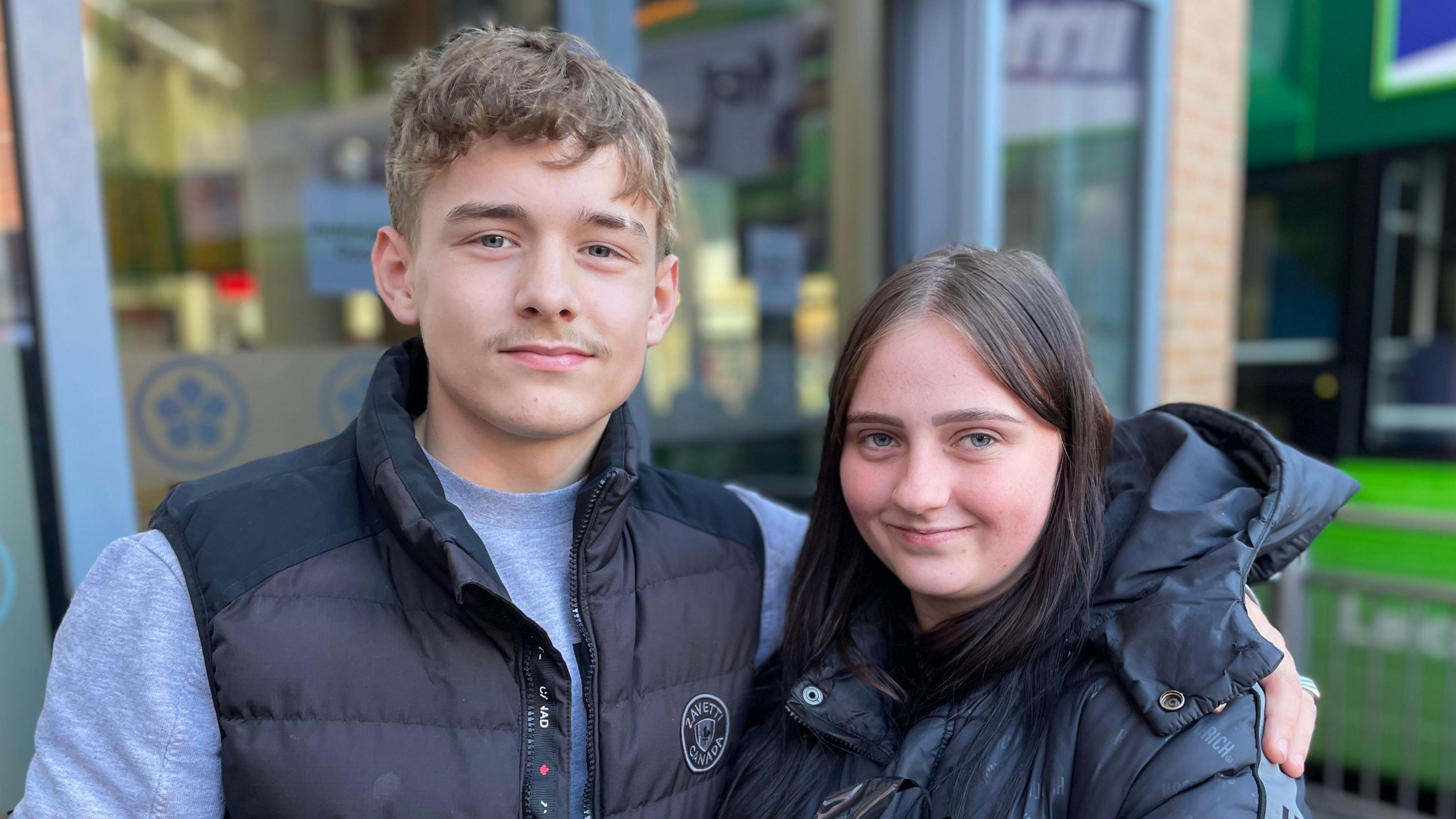 A teenage couple wearing black jackets. Kane has his arm around Amy's shoulder