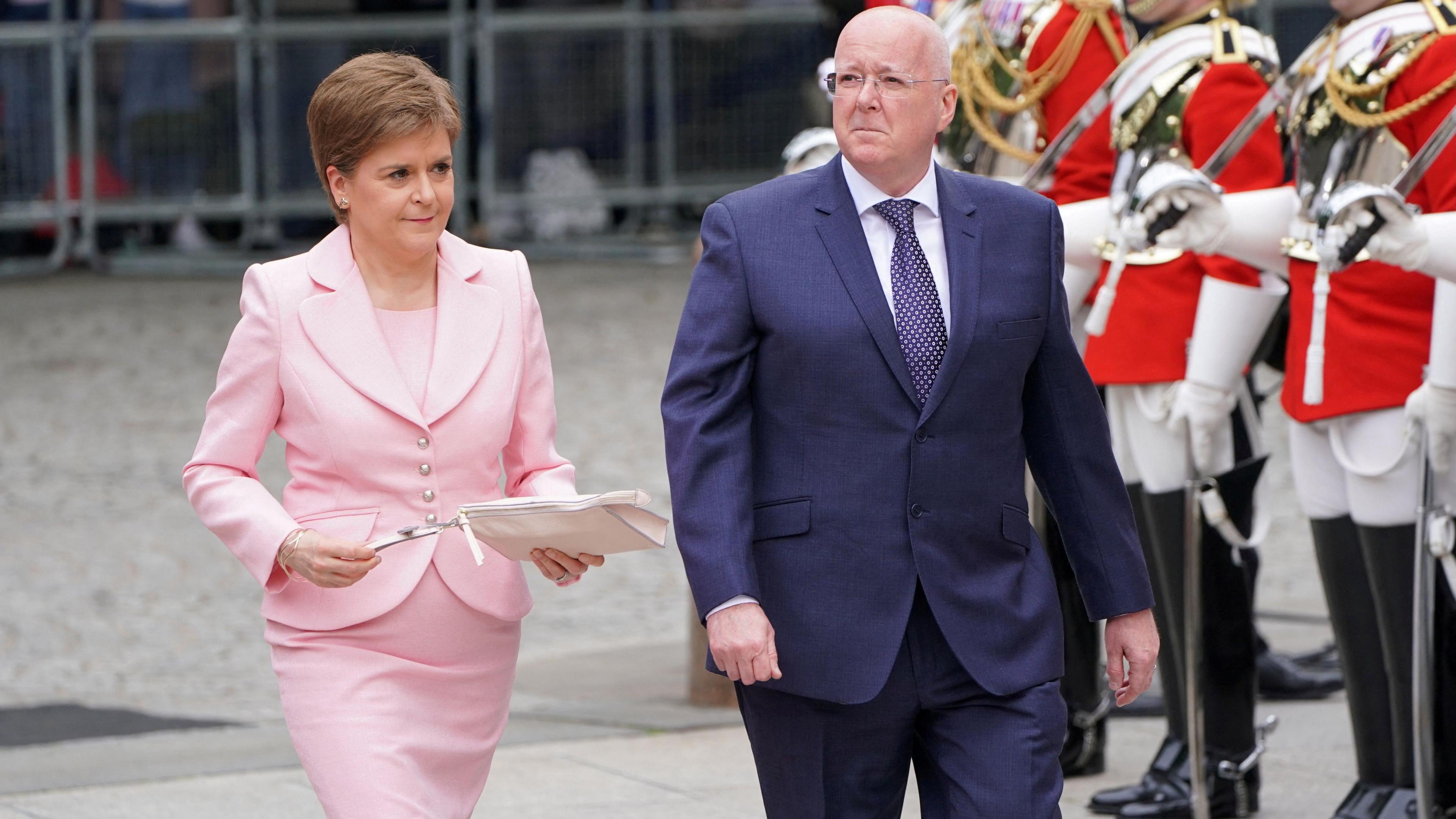 Nicola Sturgeon wearing a pink suit and Peter Murrell wearing a blue suit walk together past guards dressed in red with swords.