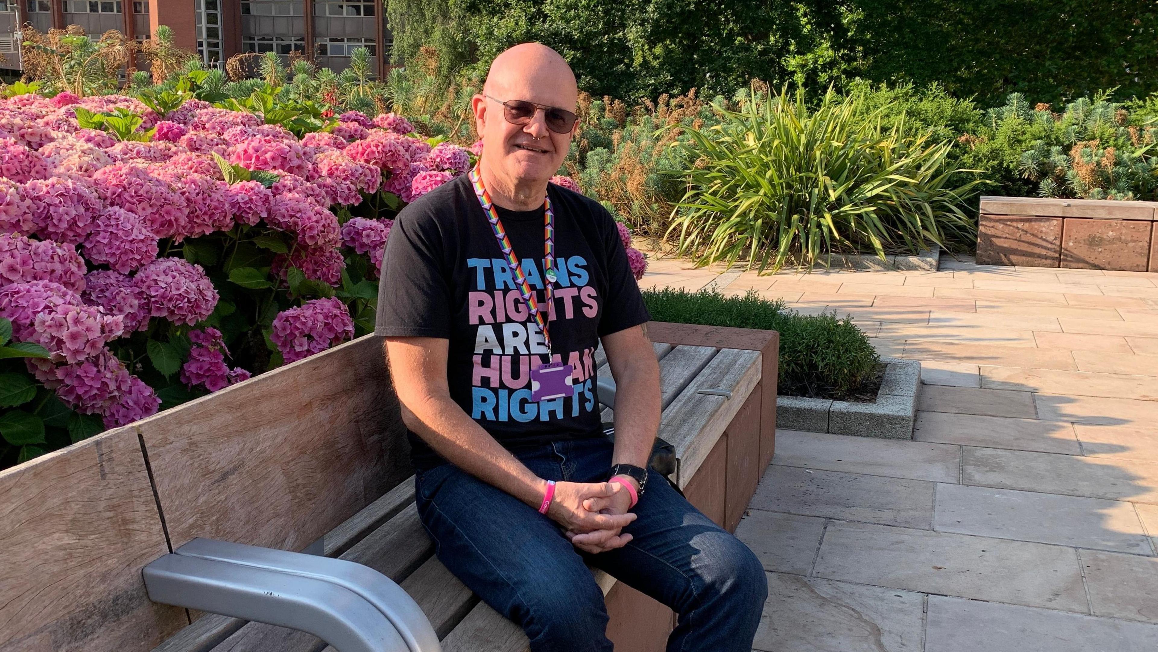 Richard Harty wearing a black top that says 'Trans rights are human rights' and sunglasses. He is sitting on a bench with pink flowers in the background