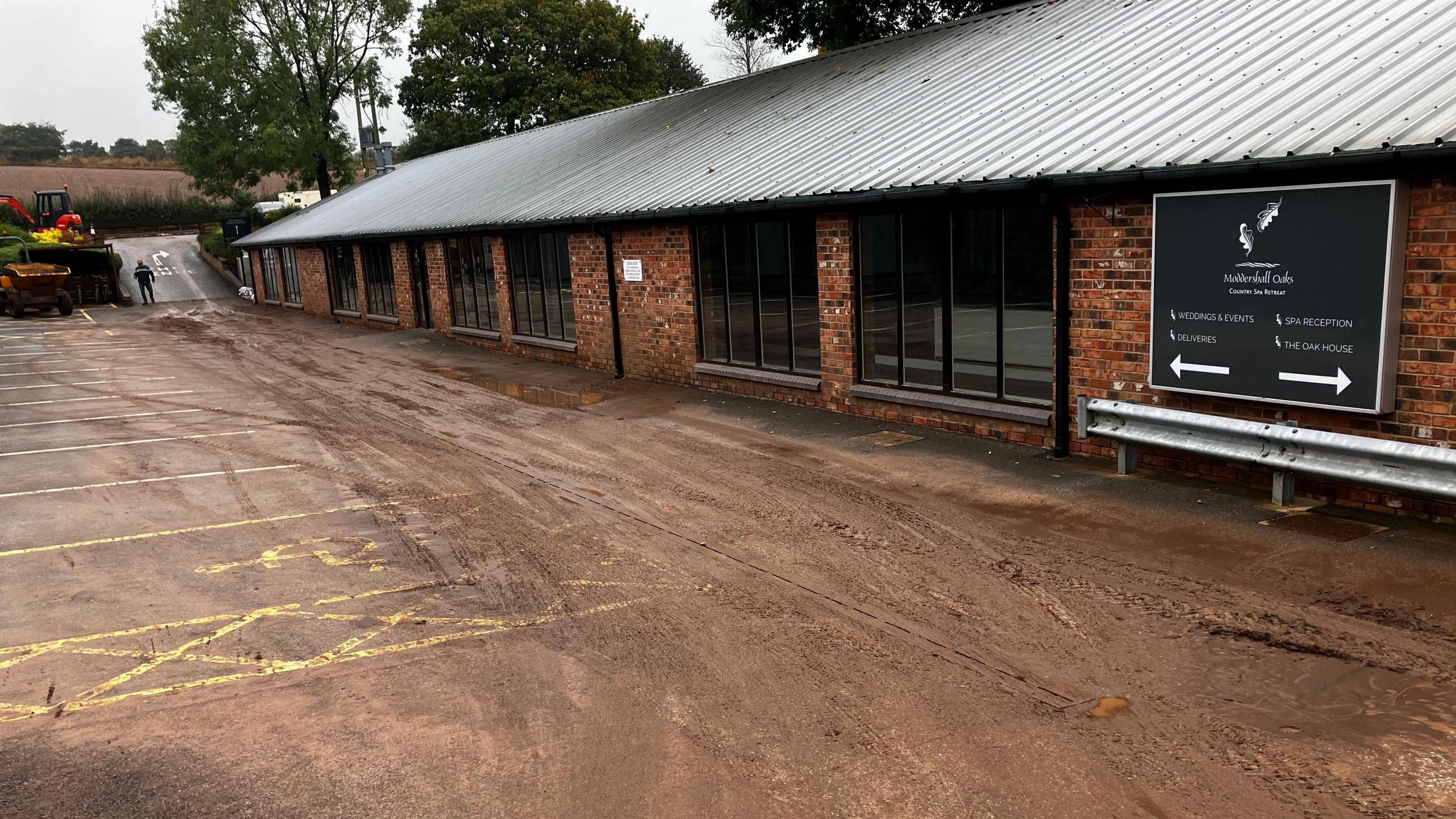 Mud and pockets of water across the ground on the car park outside a single storey brick building with a metal roof