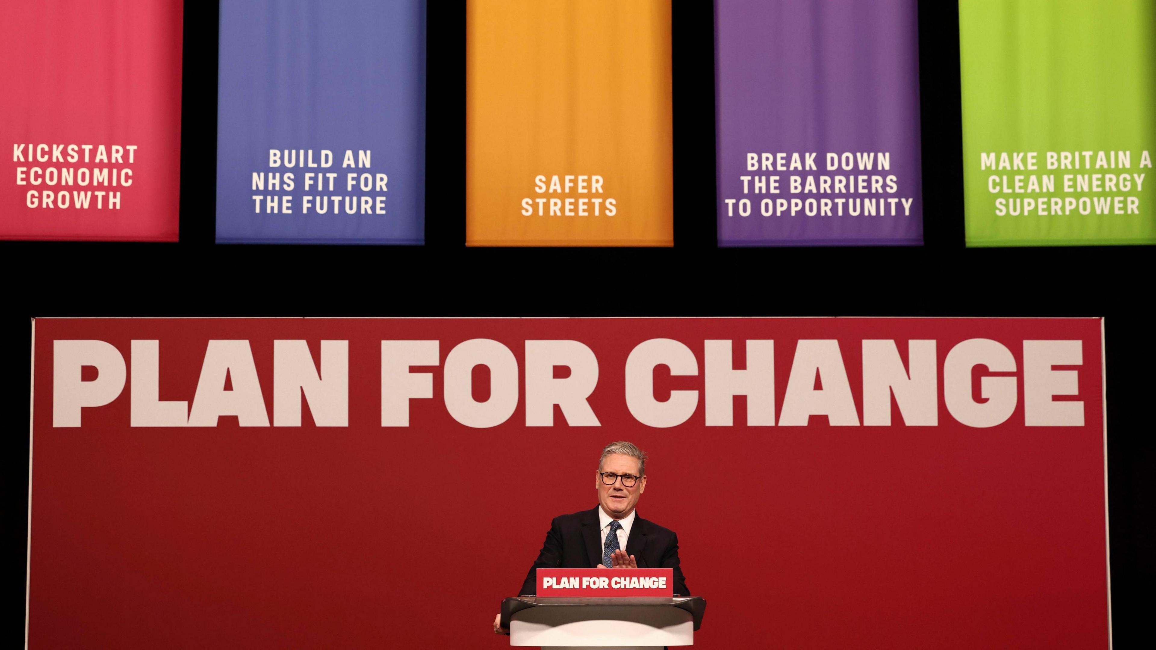 Prime Minister Sir Keir Starmer stands behind a lectern and in front of a red backdrop, both of which read 'plan for change' in white lettering. At the top of frame are five colourful banners, which read (l-r): kickstart economic growth, build an NHS fit for the future, safer streets, break down the barriers to opportunity, make Britain a clean energy superpower.