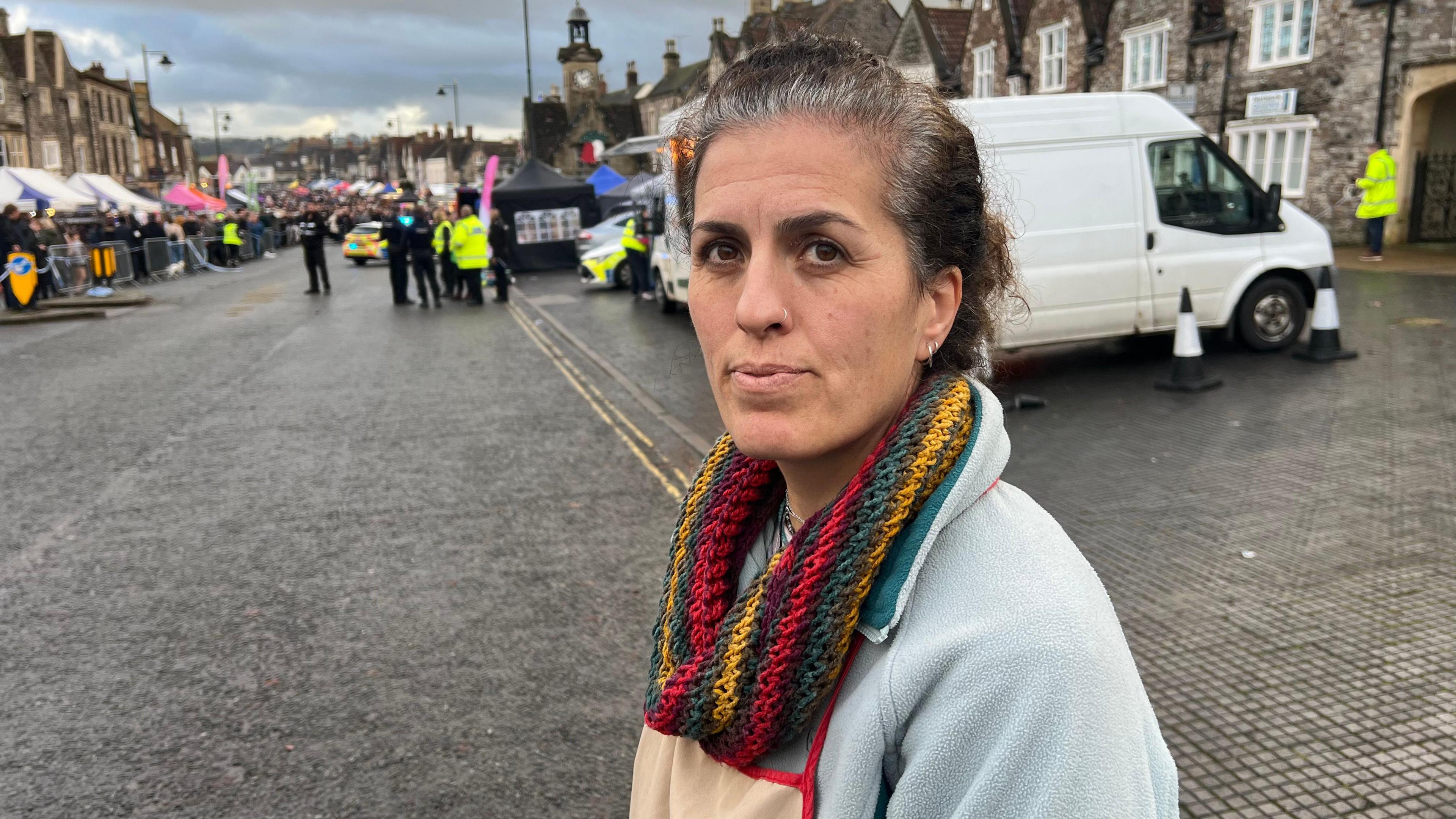 Esra Ward wearing a blue fleece pullover, a colourful knitted scarf and beige apron with red trim. She has brown hair, brown hairs and a nose ring. She is looking just above the camera, and in the background there are police gathered near the scene of the incident.