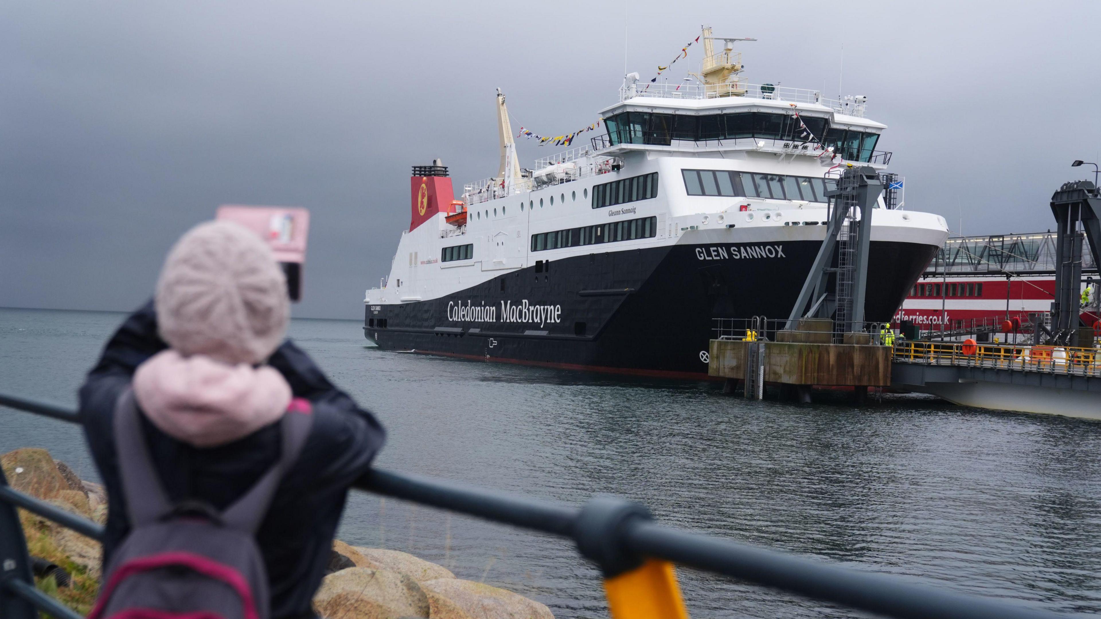 The Glen Sannox ferry arriving in Brodick on Arran from Troon on its first official day in service with the catamaran Alfred in the background. A person wearing a pink beanie hat, a grey and pink rucksack and a black jacket takes a photograph of the ferry in the foreground of the shot.