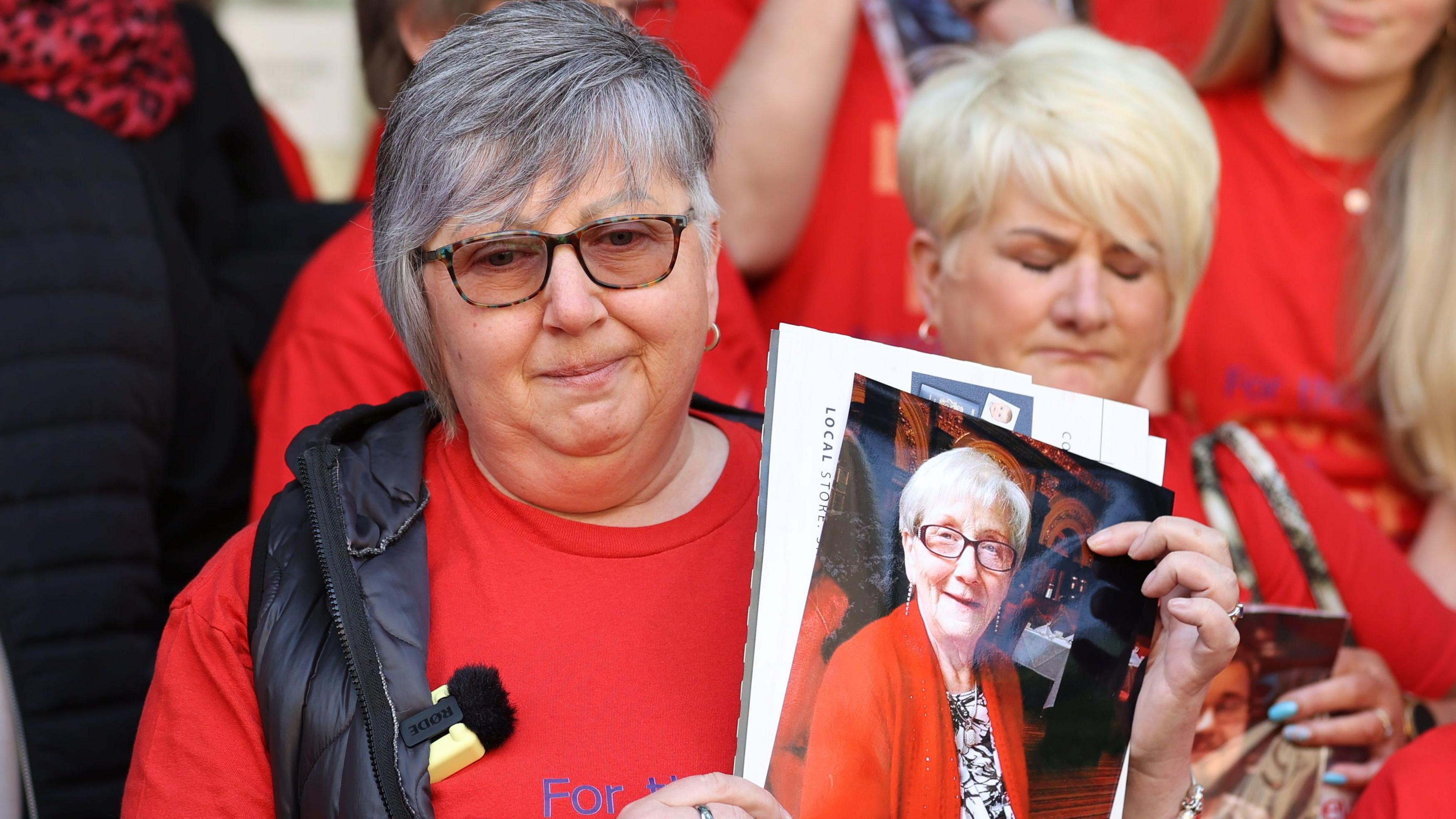 Brenda Doherty wearing sunglasses and a red top, holding a picture of her mother Ruth who is smiling wearing a red top, earrings and glasses hold a picture of her mother 