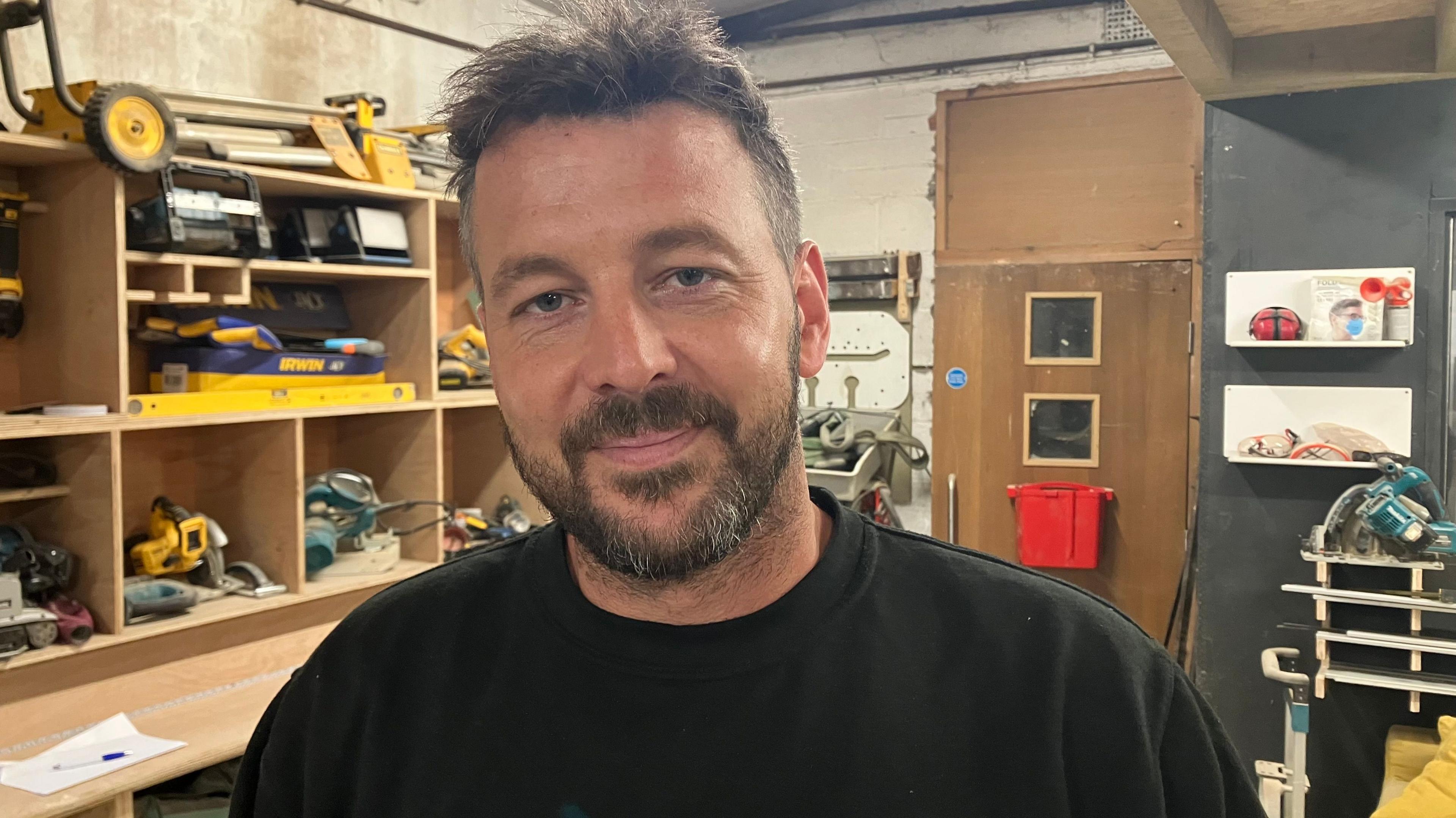Tom Popham smiling at the camera standing in his workshop. He has short, dark brown hair and stubble. There are wooden shelves and cubby spaces behind him filled with tools.