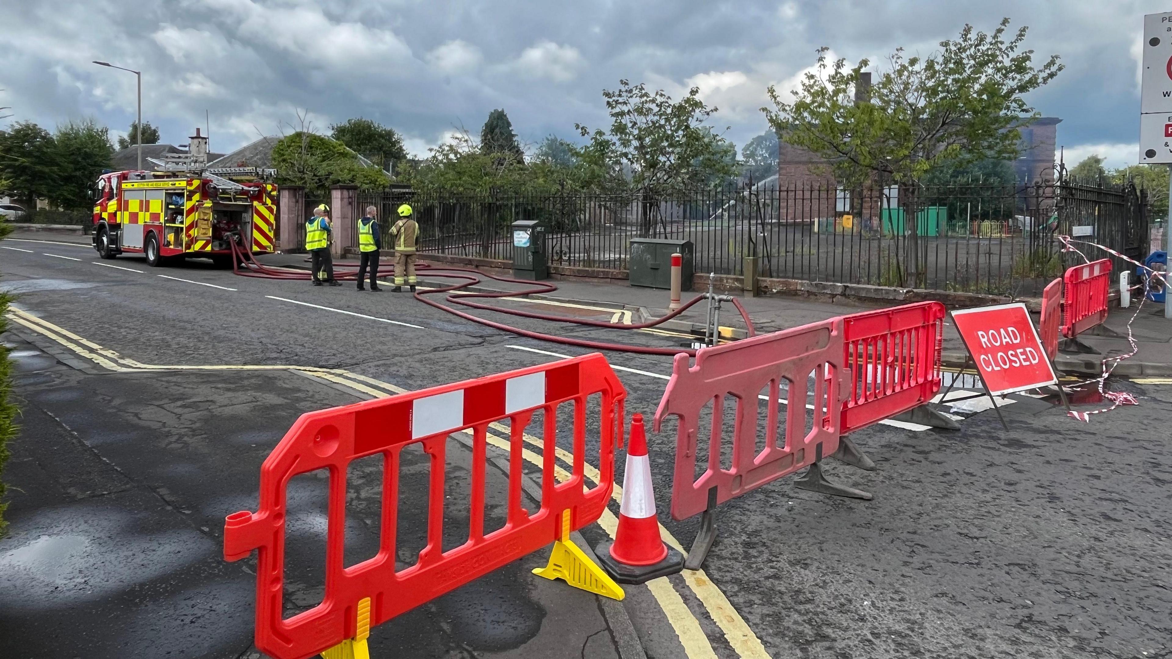 Fire crews spraying water at the school building. There is a red plastic fence in the foreground of the picture.