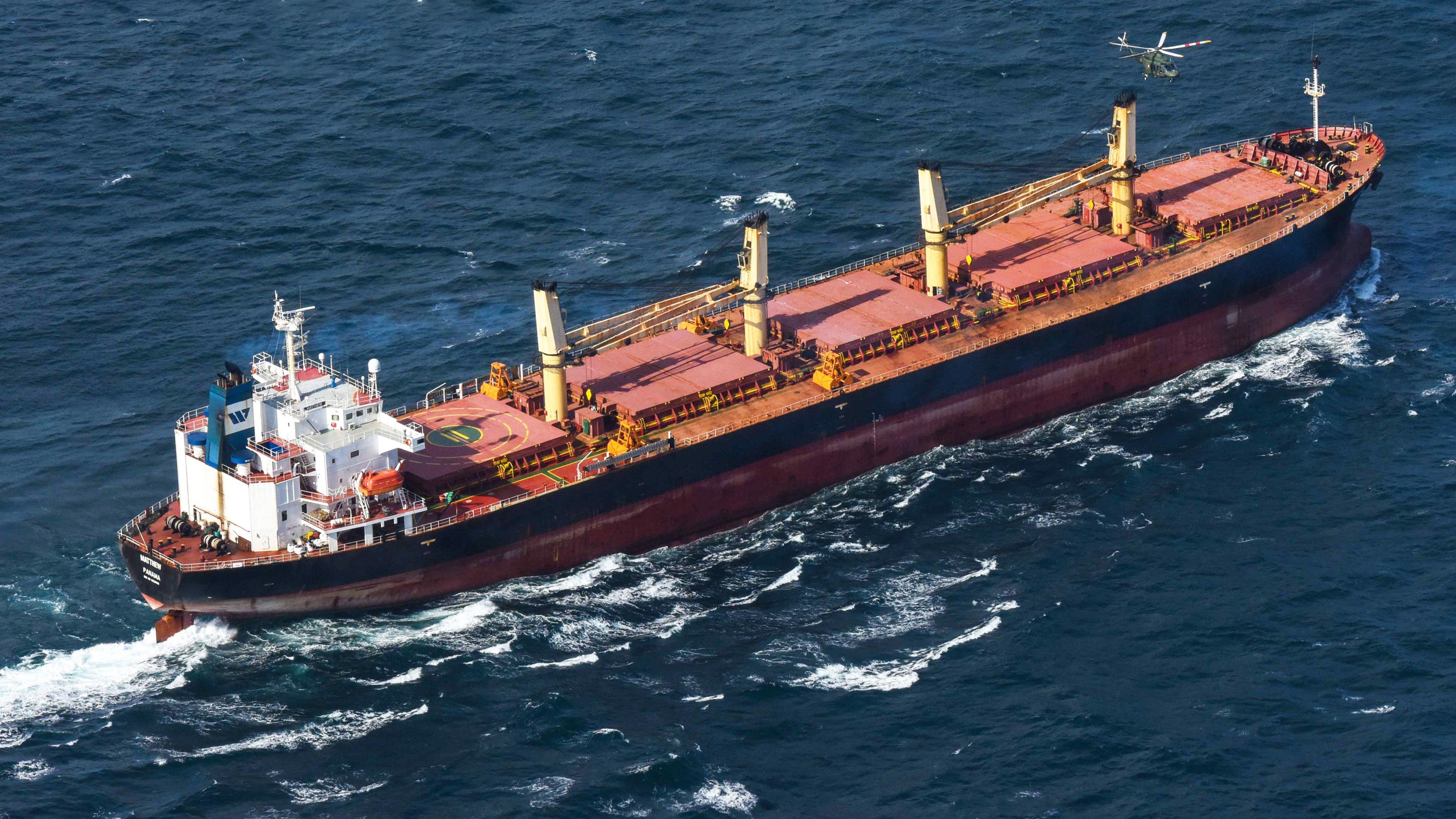 A large red-painted ship is at sea with a helicopter hovering above it. The ship is moving through the water causing a stream of white waving water behind it. The ship has a large white painted wheel house, four large chimneys and a blue strip painted around it's exterior.
