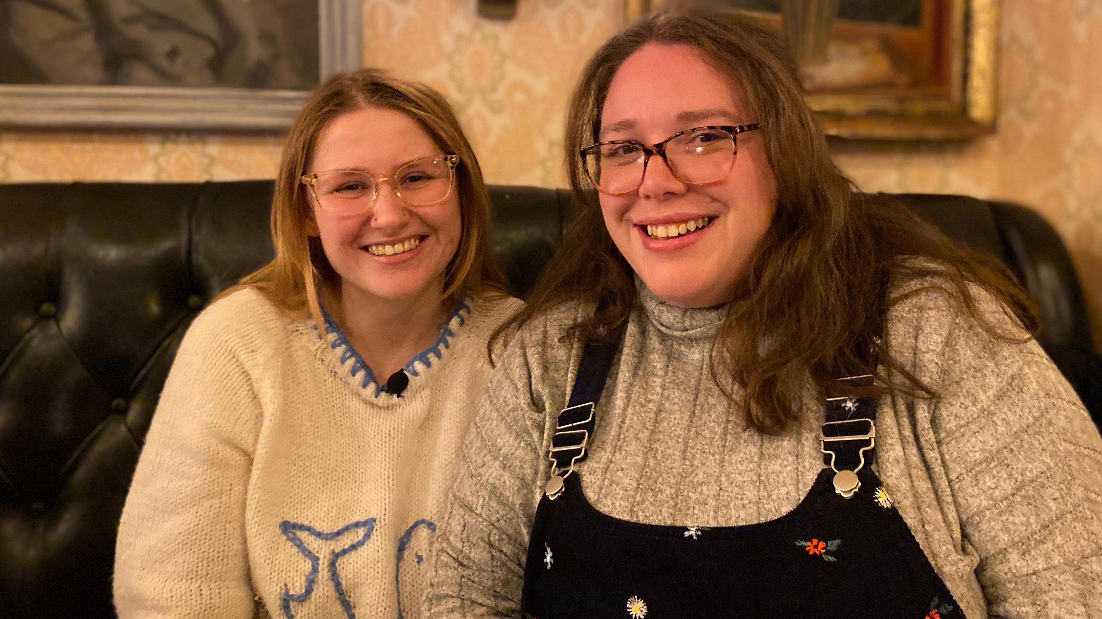 Lo Ranfaing is smiling with Hollie Boulton. Lo has long brown hair, is wearing glasses and a white and blue jumper. Hollie has brown hair, is wearing glasses, a grey jumper and a black pinafore. They are sitting on a brown leather sofa.