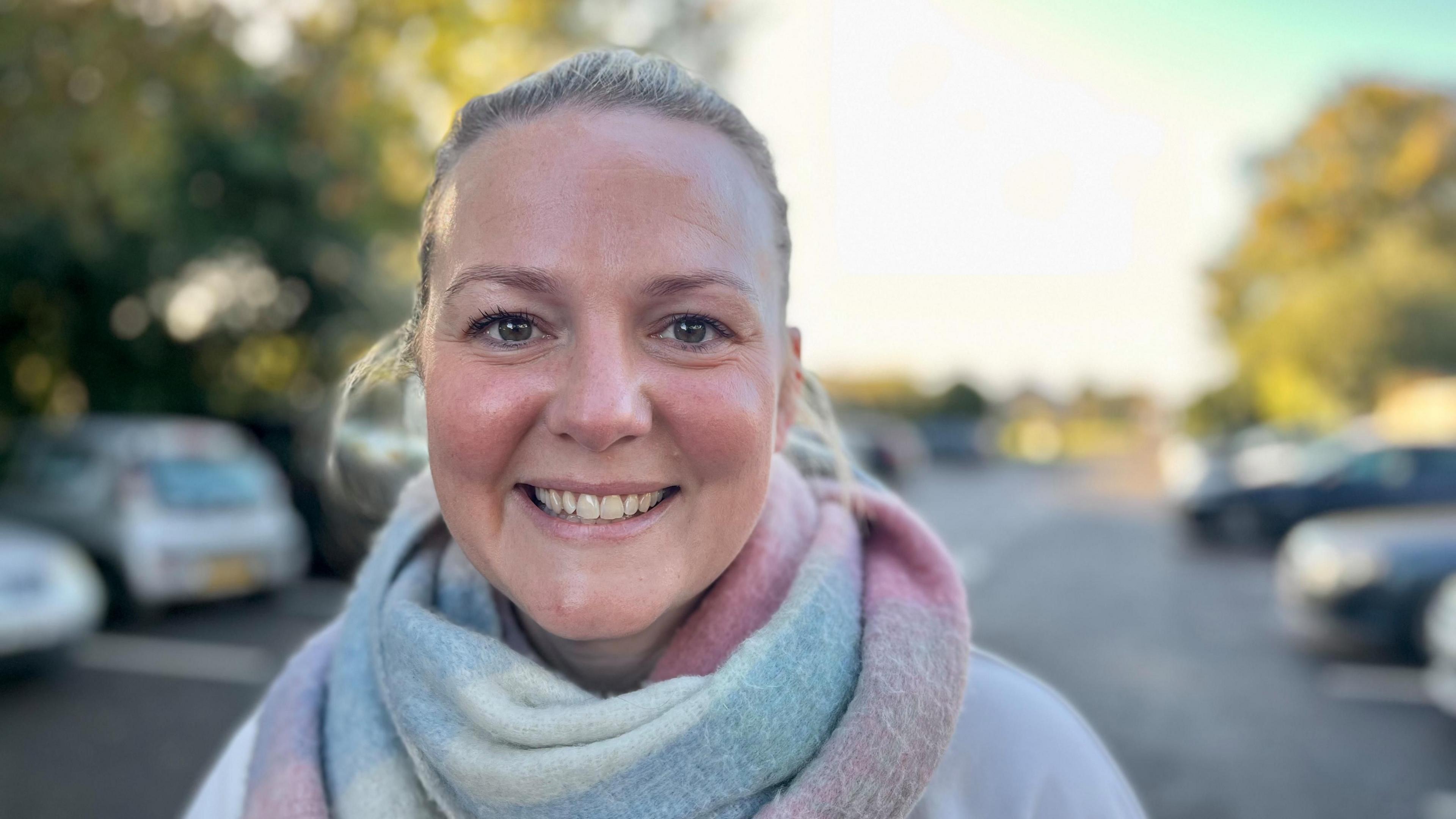 Charlotte Reed has long, blonde hair tied in a pony tail. She is smiling at the camera, wearing a cream jumper and a pink, blue and cream scarf wrapped around her neck. Behind her there are parked cars in the Butts Lane car park