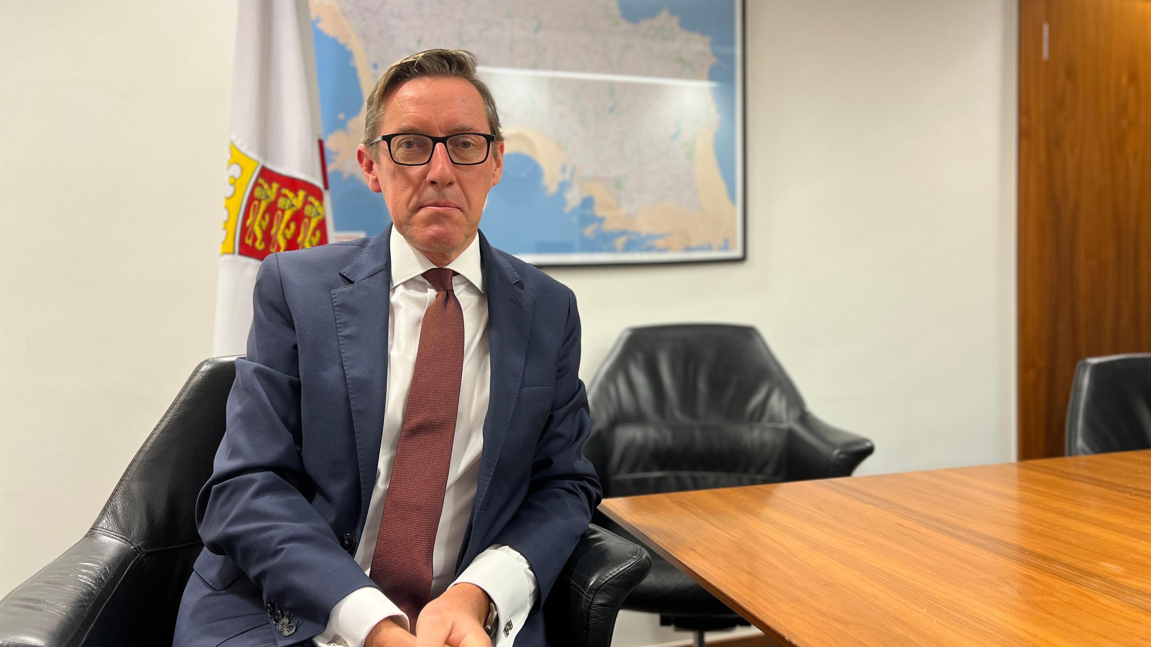 Ian Gorst looks at the camera as he's sat on a black leather chair. Behind him is a Jersey flag and a map of Jersey on the wall as well as more black chairs. There's a conference room table next to him. He's wearing a navy blue suit with a light red tie with white dots. He has glasses with dark frames which are rectangular.