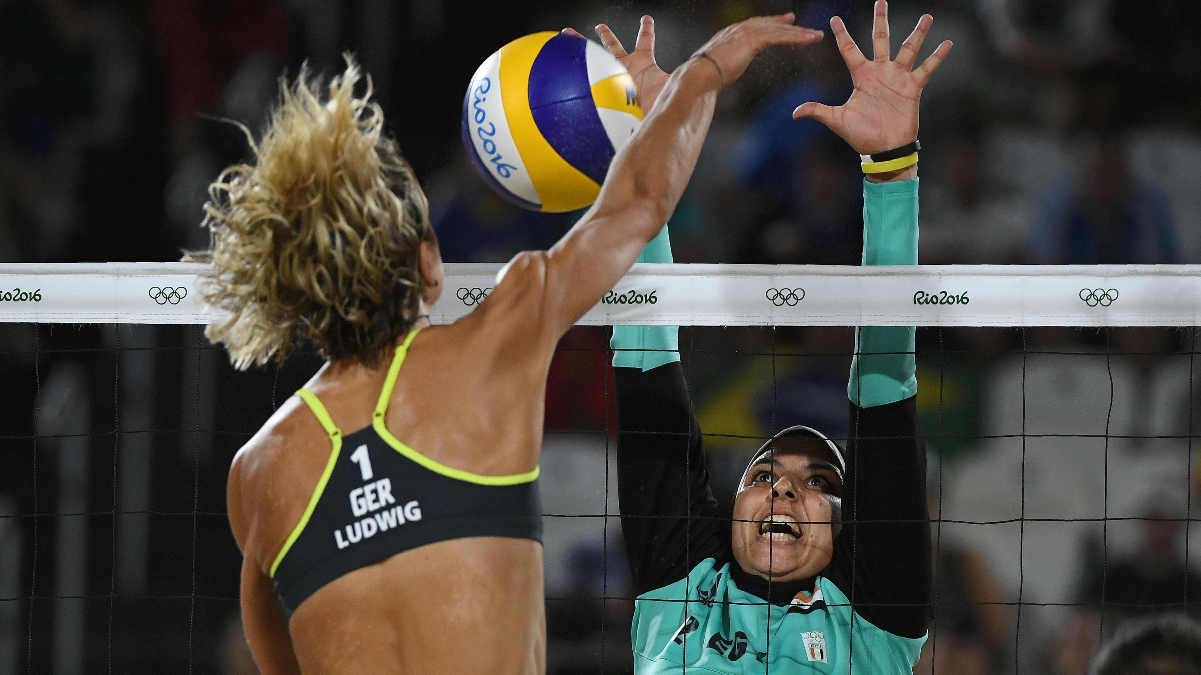 Doaa Elghobashy in Egypt kit with long black sleeves and headscarf jumps to try to block a spike at the net in a 2016 Olympic match against Germany as her opponent, in a bikini is on the near side of the net