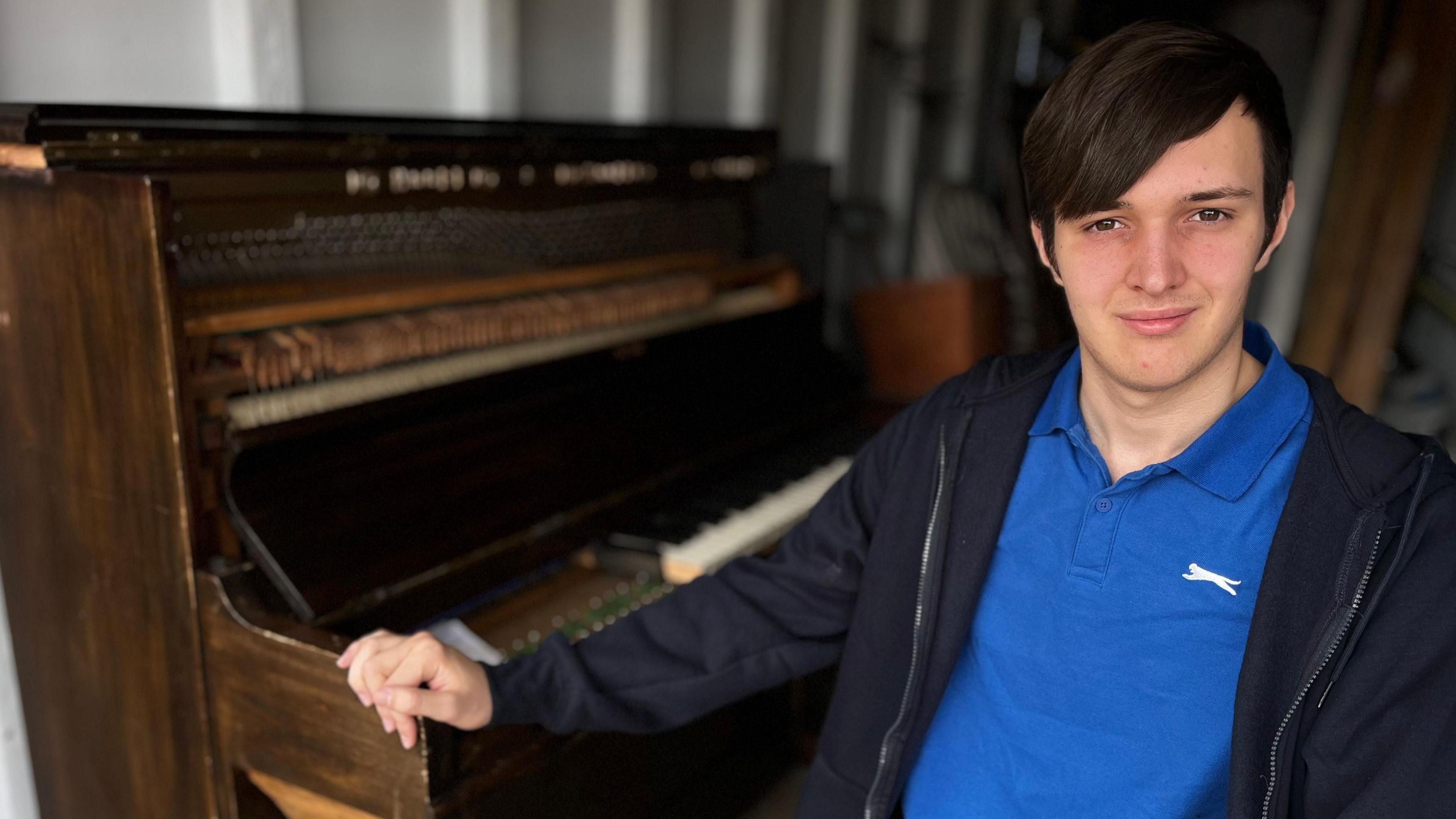 Jamie is leaning on the wooden piano. He is wearing a blue polo shirt with a navy blue hooded jumper over the top. He has dark brown hair, with his fringe swept to one side. He is looking at the camera.