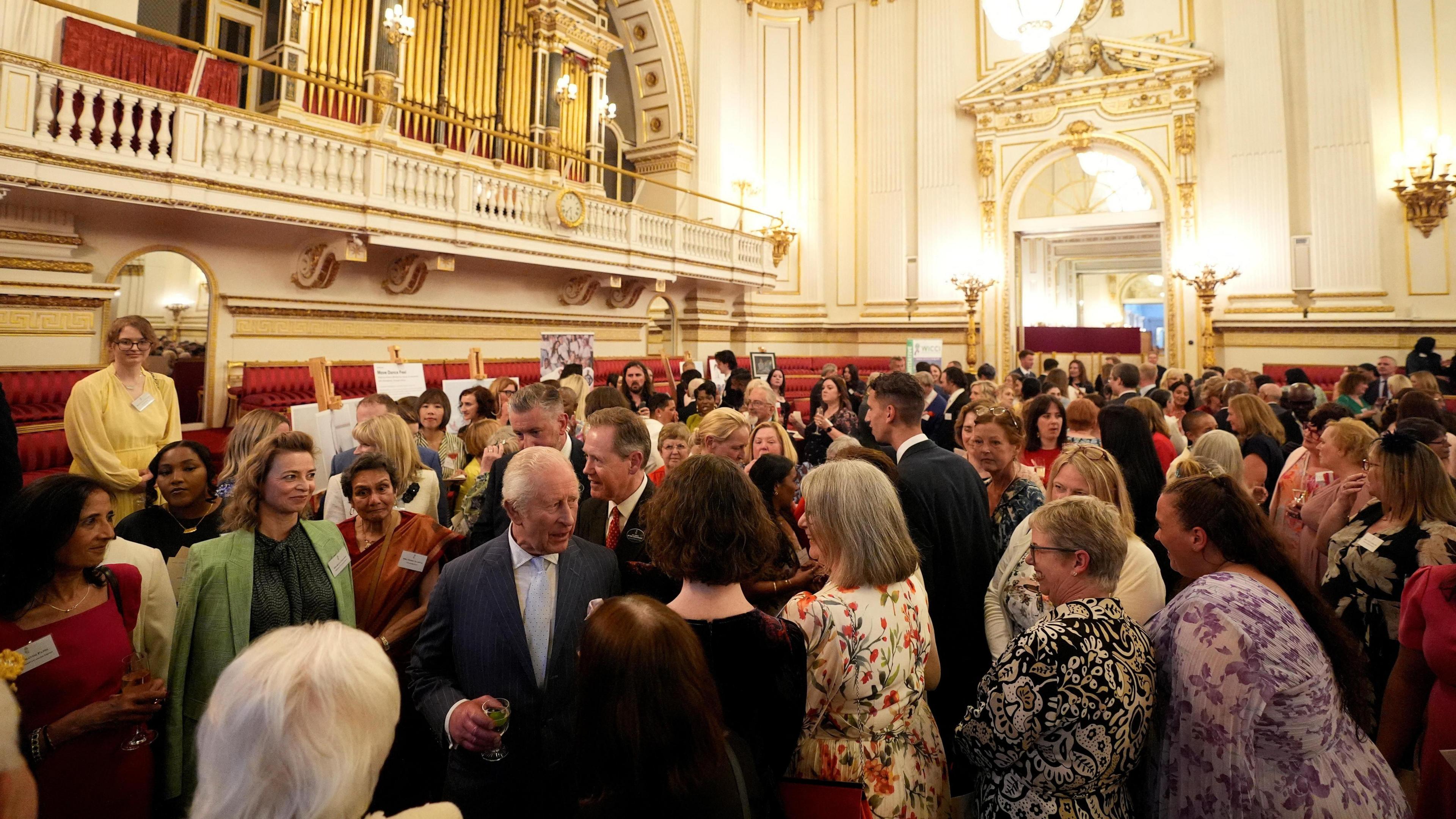 A huge crowd of people in an ornately-decorated room with what looks like a pipe organ on a balcony. King Charles is in the midst of the crowd with a drink in his hand as he talks to people.