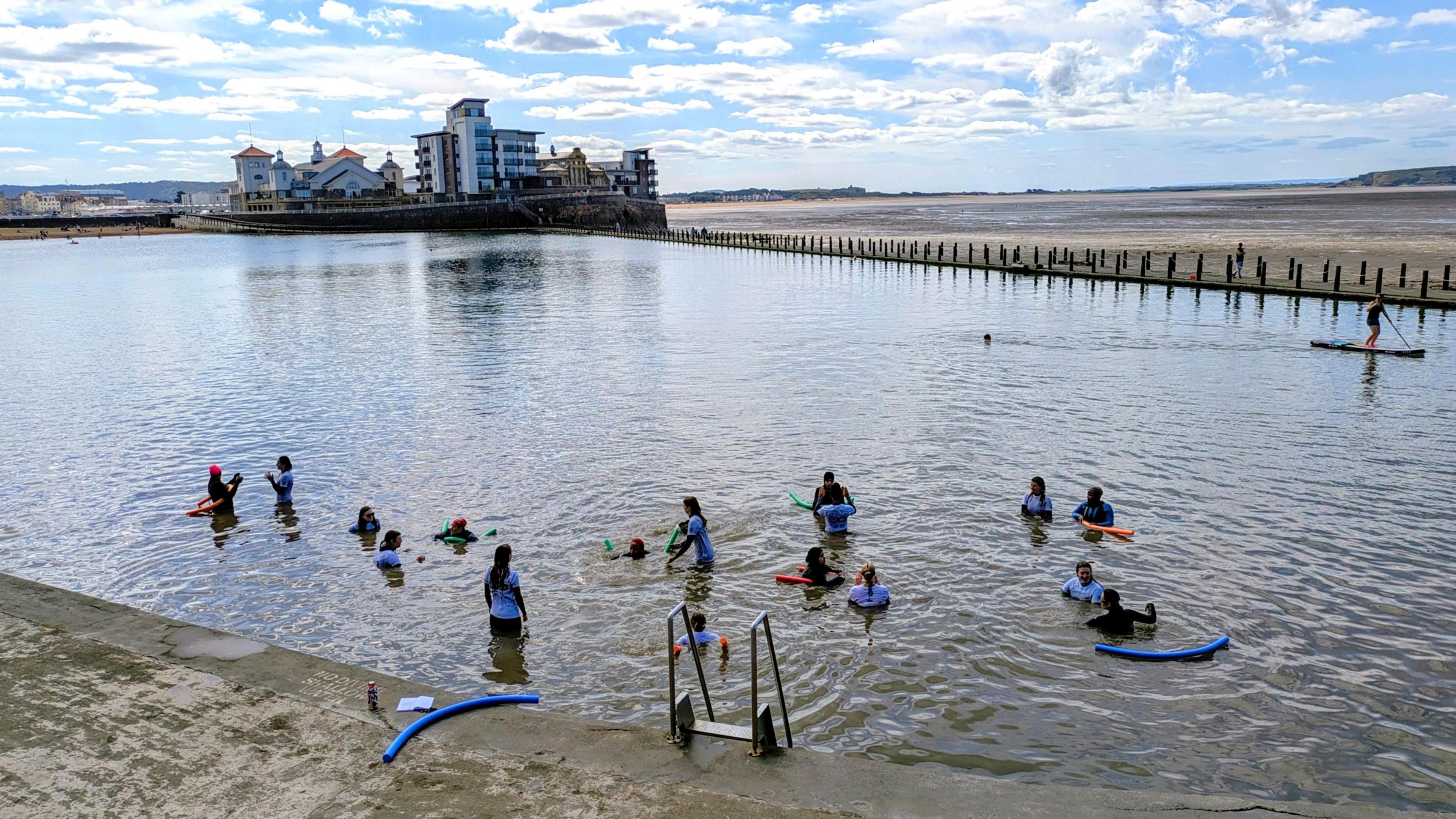 A group of eight women and their instructors waist deep in Weston-super-Mare's Marine Lake. They are wearing white rash-vests and have floatation devices to help them learn to swim 