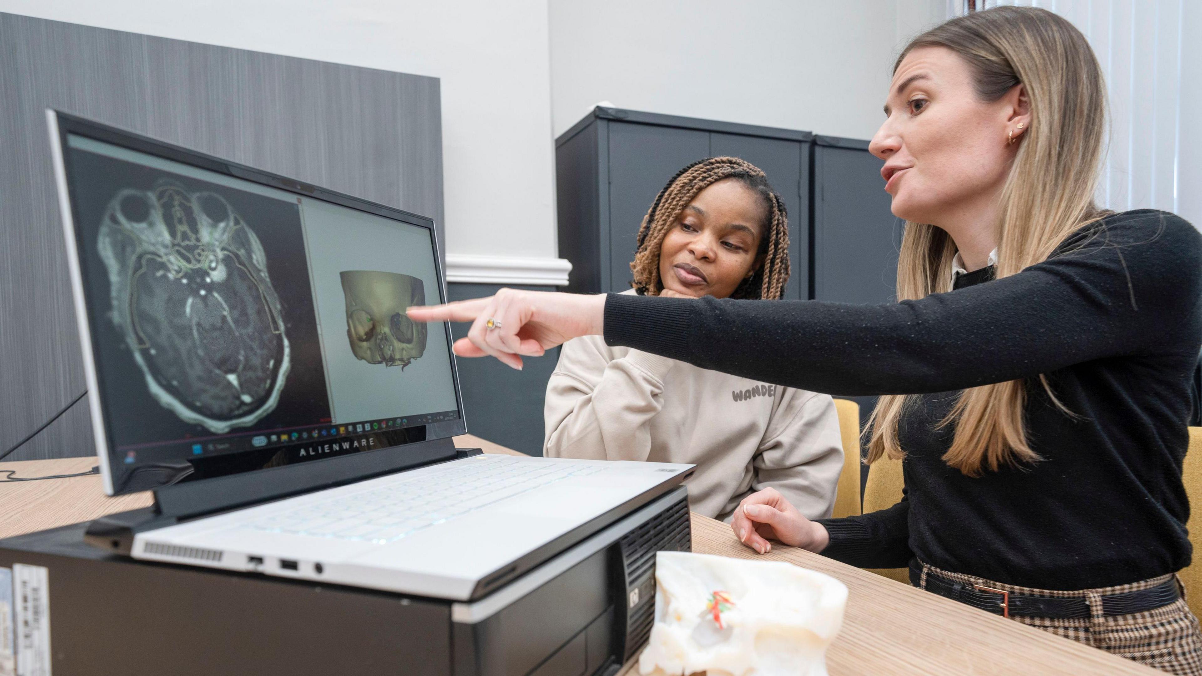 A woman with long fair hair and a black jumper points to a model of Ms Kaviya's skull on a computer screen. Ms Kaviya sits alongside her looking thoughtful.