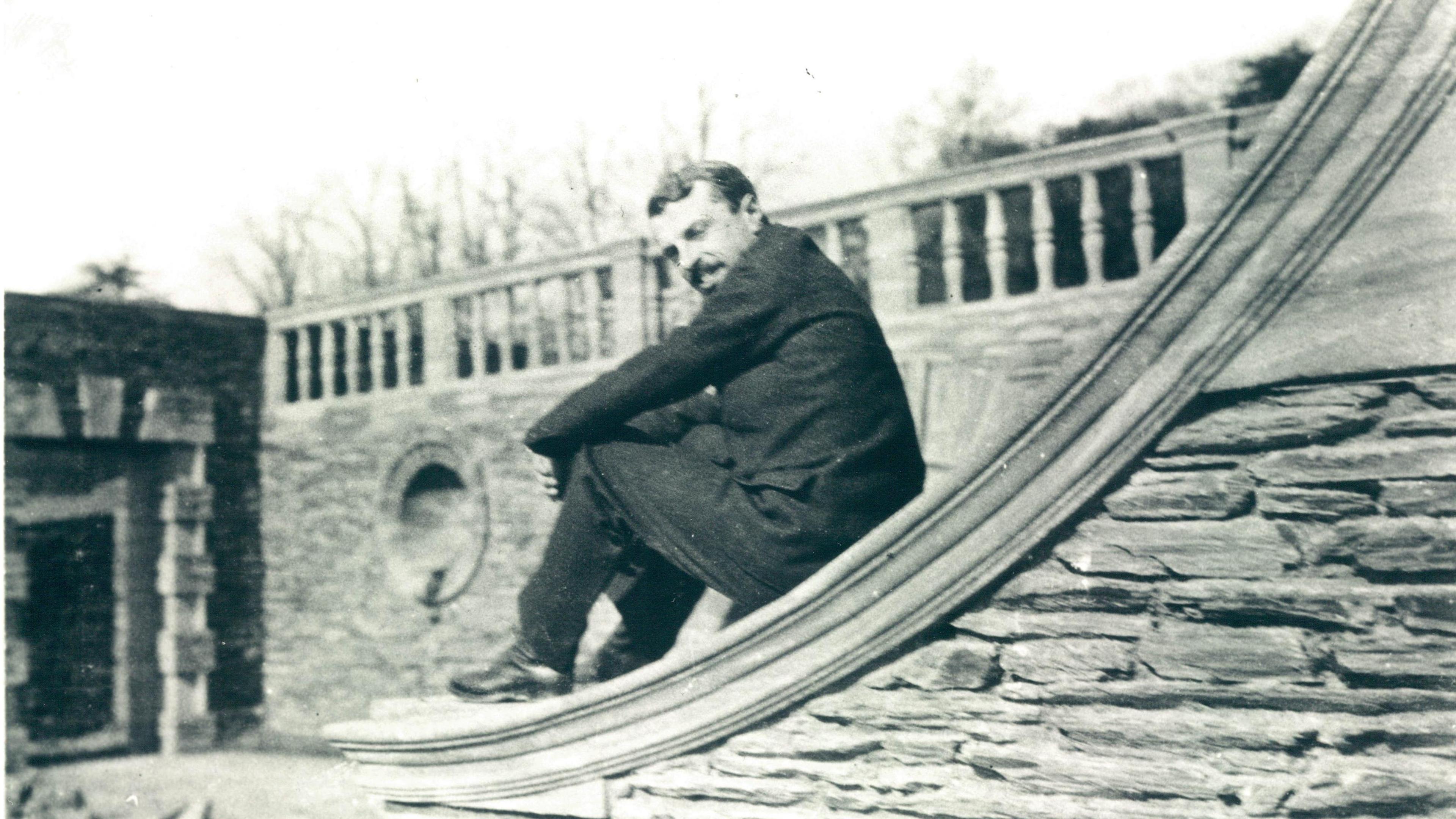 A man in a dark suit sits on the bottom of a curved stone wall. In the background is a stone banister on top of another stone wall.