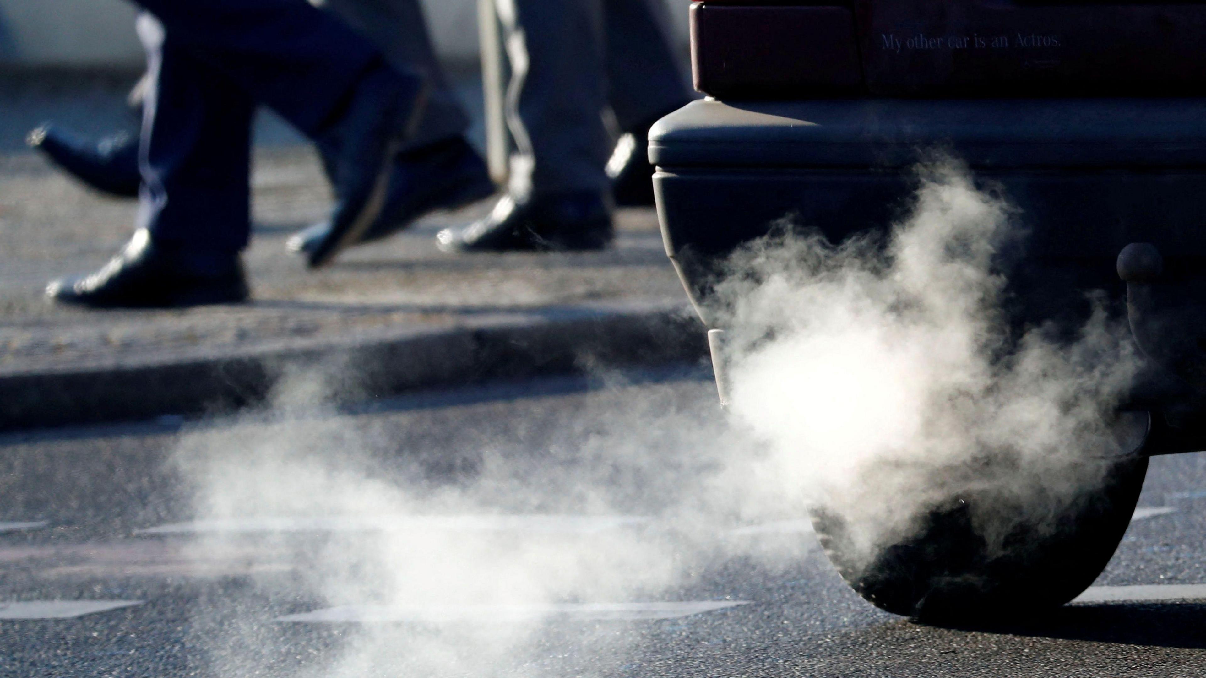 White smoke emerges from a car exhaust on a street. Three men in suit trousers walk by on a pavement in the background.