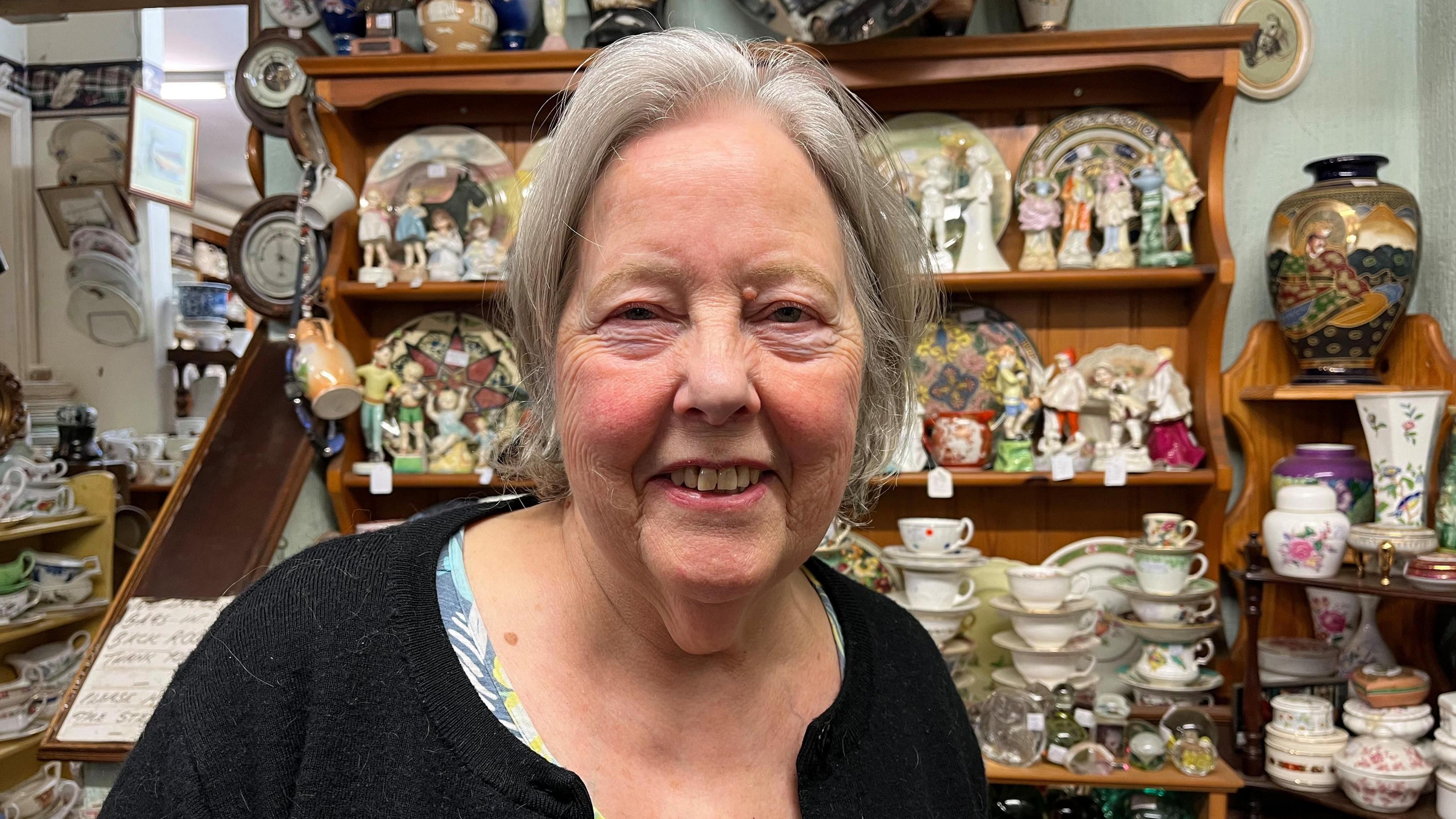 A woman with tied-back grey hair smiles at the camera. She is wearing a black cardigan and standing in front of shelves covered with cups, plates and ornaments.