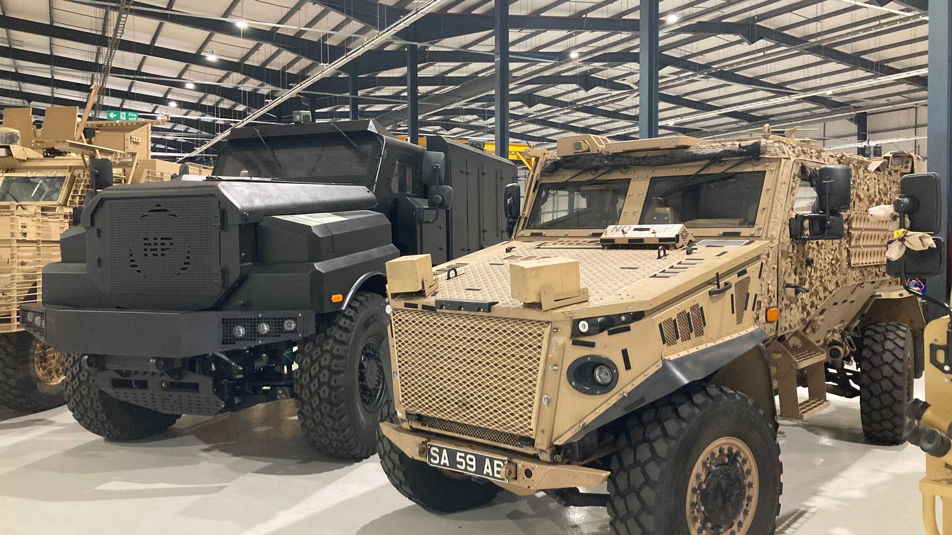 A black Foxhound vehicle and a beige Ridgeback vehicle sit site by side in the factory. The factory has a metal, ridged roof which is grey overhead.