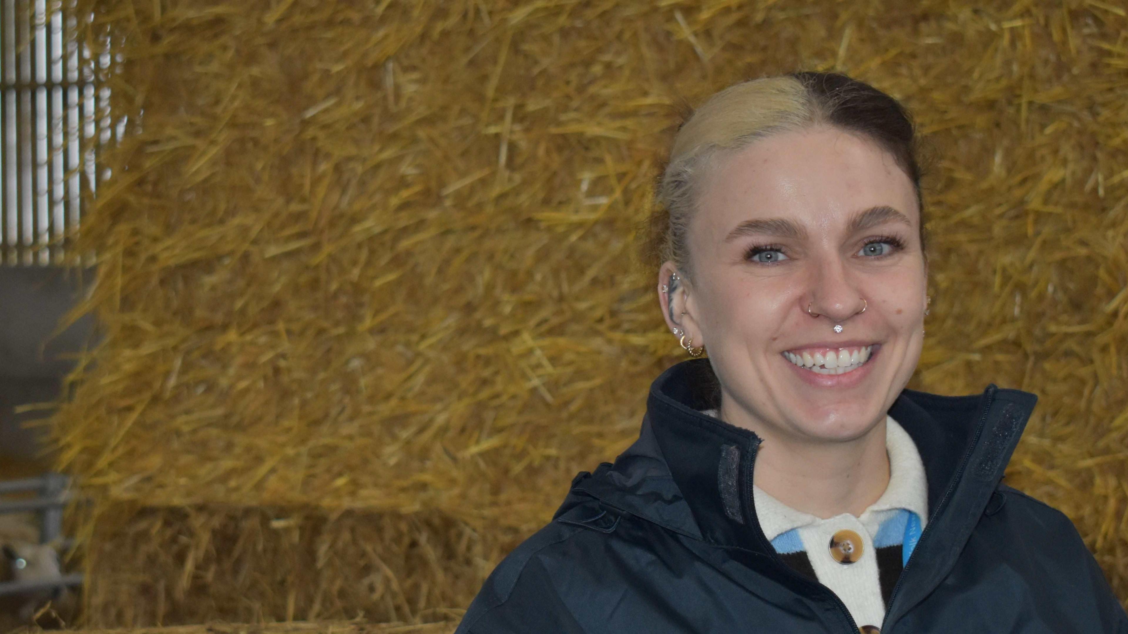 Abbie stands in front of a large straw bale. She is wearing a dark blue mac with the collar up and a white rugby shirt collar visible underneath. Her hair which is half blonde half brown is tied back