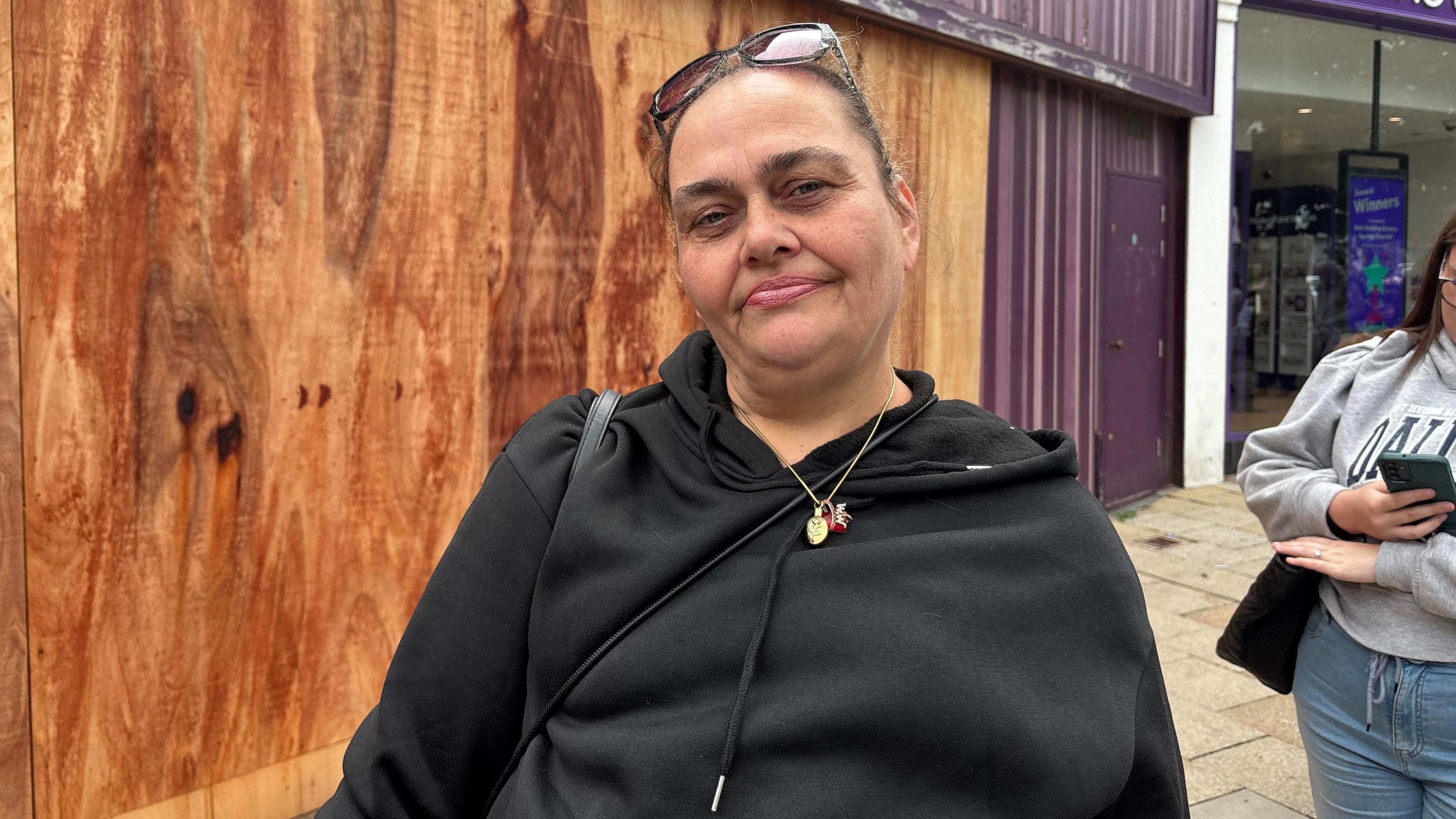 Tracey Johnson is wearing a black top and gold necklace and she has sunglasses on her head. She is standing in the queue for the shop. Behind her, another business is boarded up. Another shopper, who is looking at her phone, can be seen behind her.