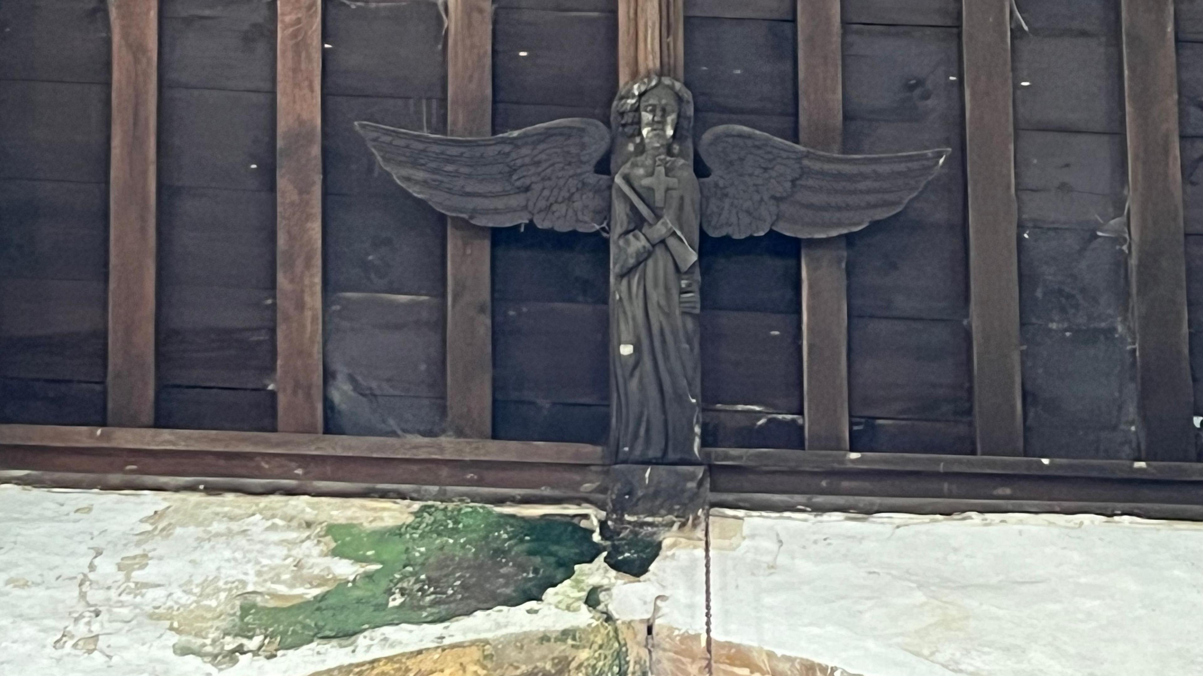 Carved wooden angel around 1m (3ft) long and a similar width fixed at the top of a wooden ceiling in a church, green moss or mould can be seen at the feet of the angel which is caused by a leak in the roof.