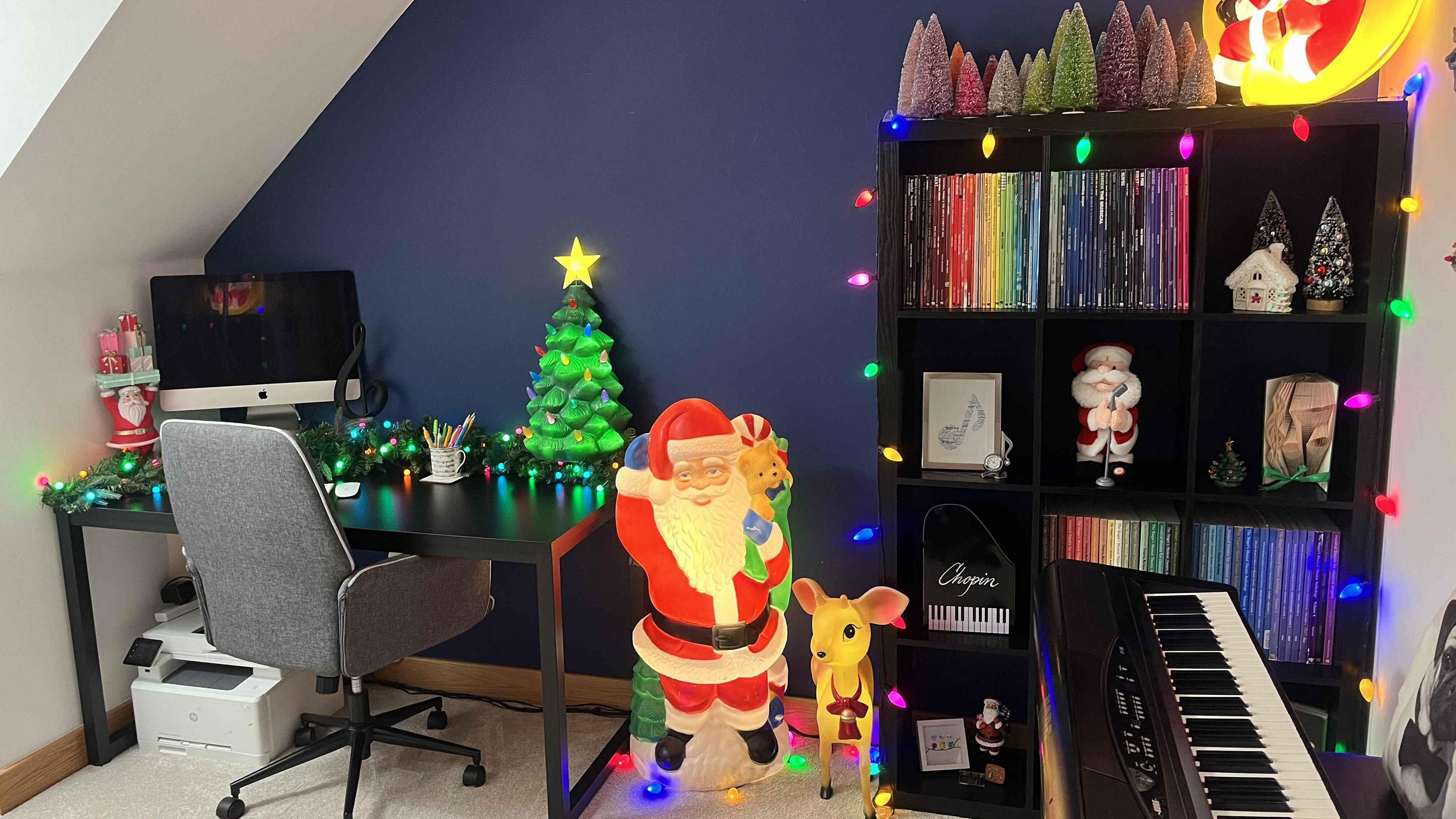 An office full of festive decorations. The desk is bordered with a garland interlaced with multi-coloured fairy lights. A light up Santa with a sack full of toys and a small light up reindeer stand next to the desk. A bookcase, which contains vinyl records arranged by colour, is bordered by more multi-coloured lights and a row of small colourful Christmas trees sit on top.