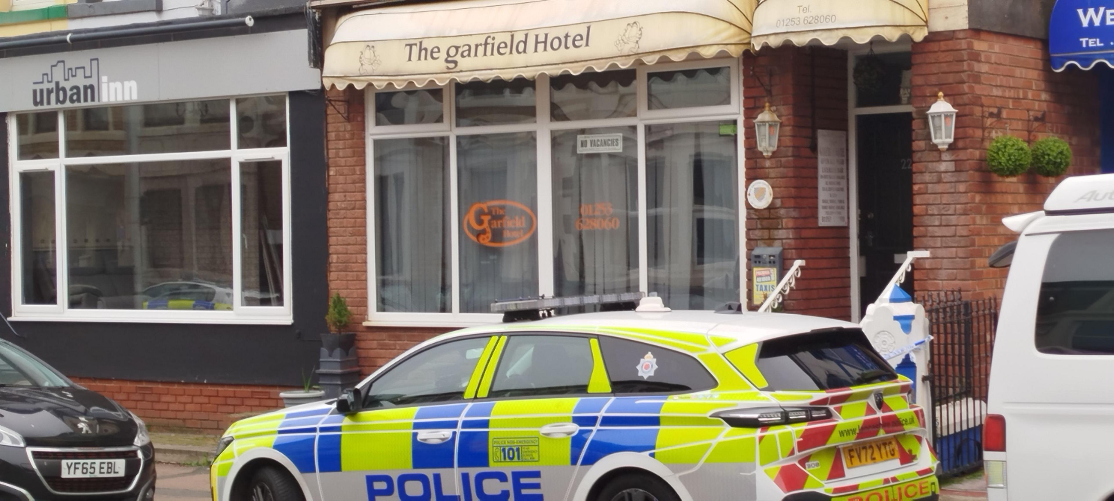 Police car outside Garfield Hotel- a red brick building with cream frilled canopy. The orange writing on window reads Garfield Hotel. A sign says No Vacancies. 