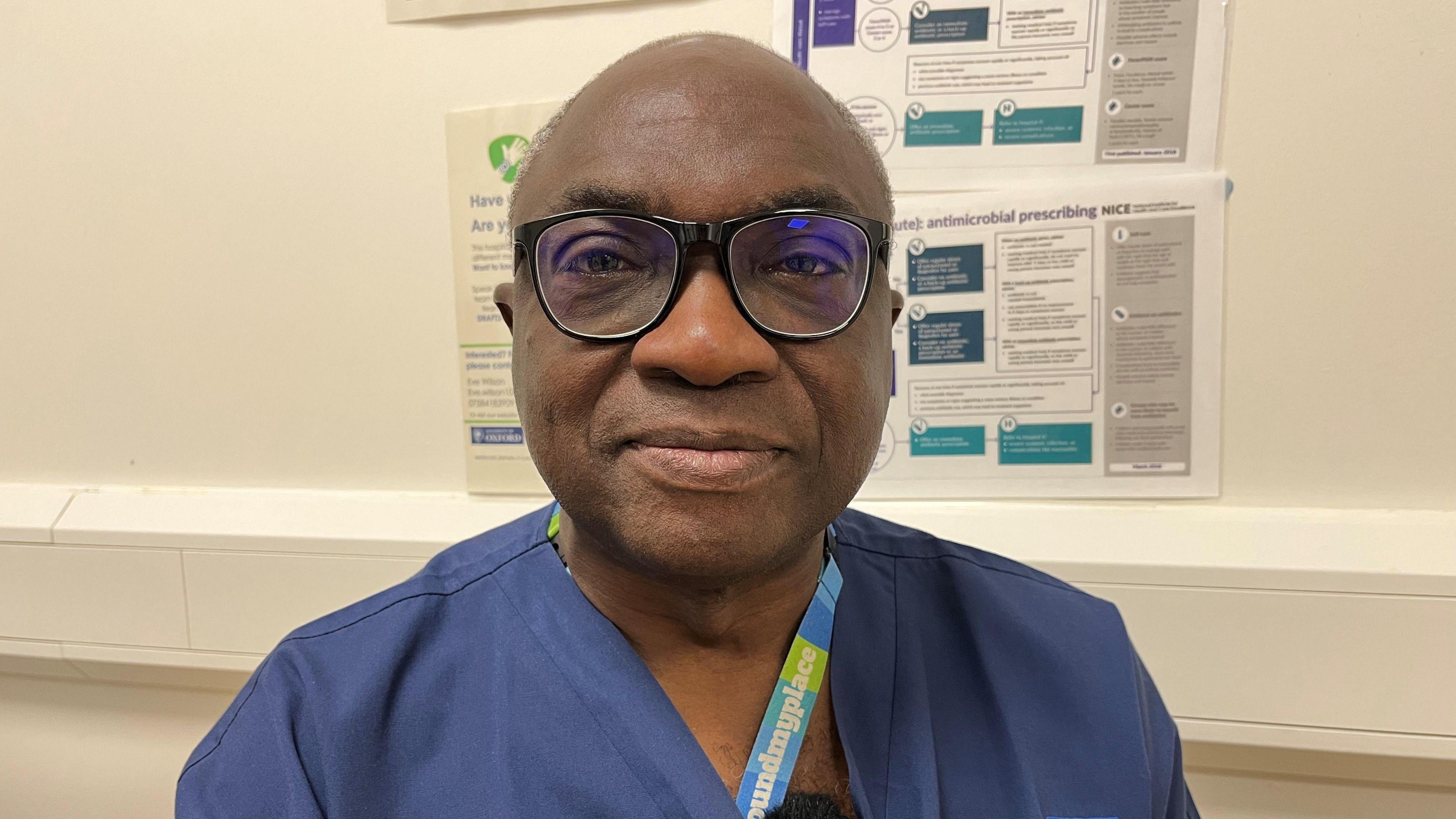 A man in his 40s with glasses staring into the camera, wearing scrubs.