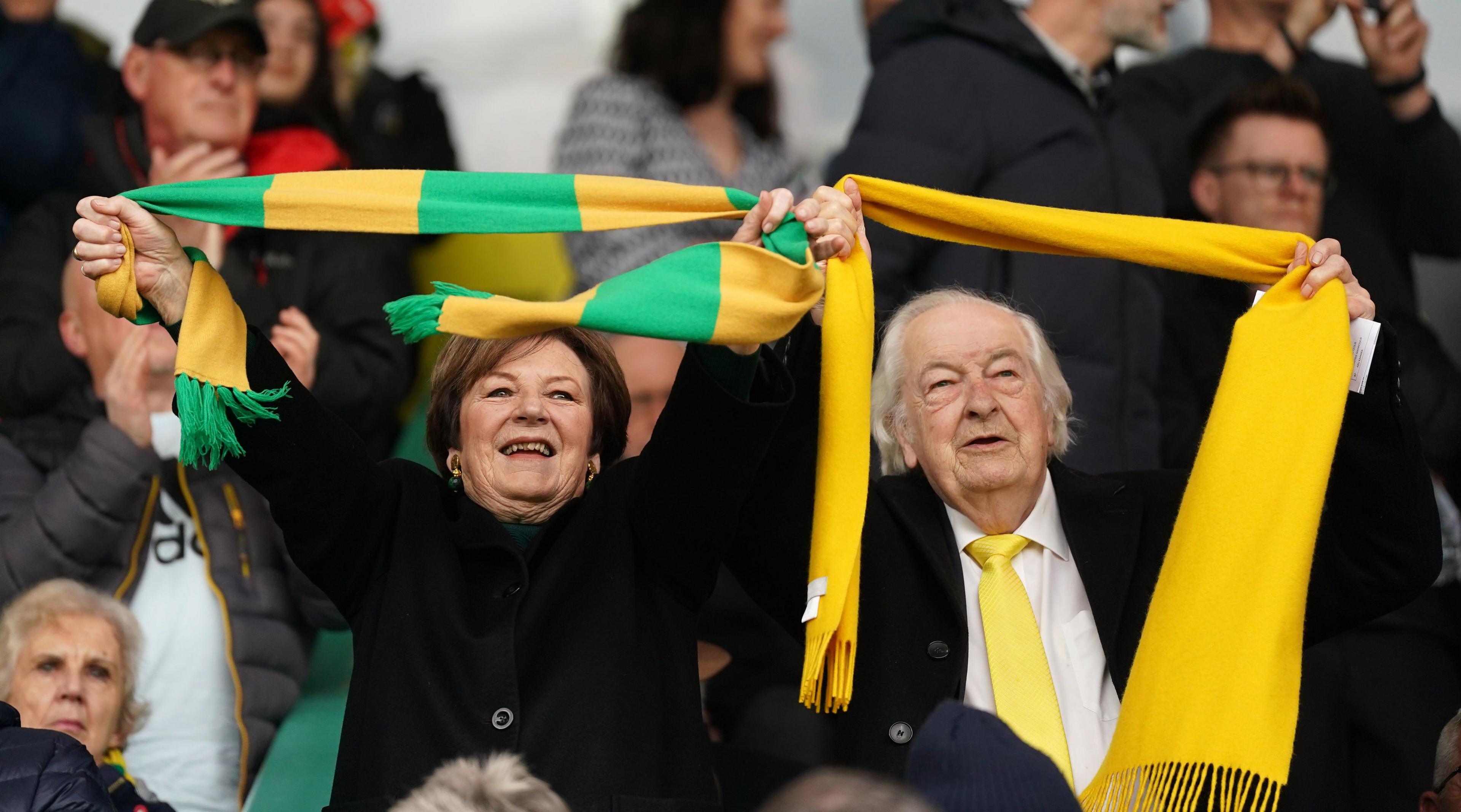 Norwich City directors Delia Smith, left, and Michael Wynn Jones cheer on their team during a Championship match at Carrow Road in April 2023. Delia is wearing a black jacket and holding a yellow and green scarf. Michael Wynn Jones is wearing a black coat and a white shirt and yellow tie. He is holding a yellow scarf. They are in front of a group of people.