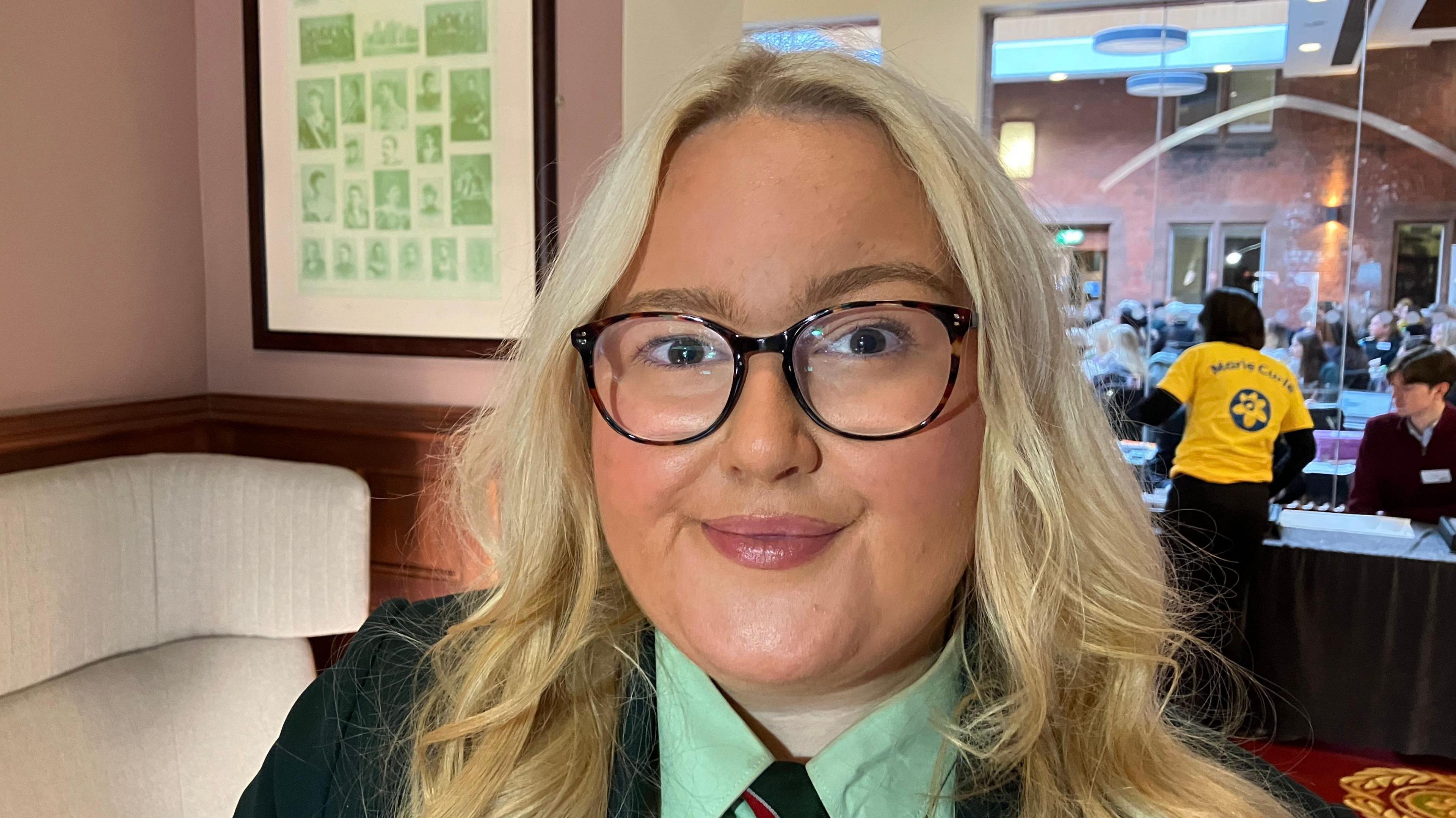 Darcy has long blonde hair with brown eyes. She is wearing dark glasses and is wearing a green school uniform.