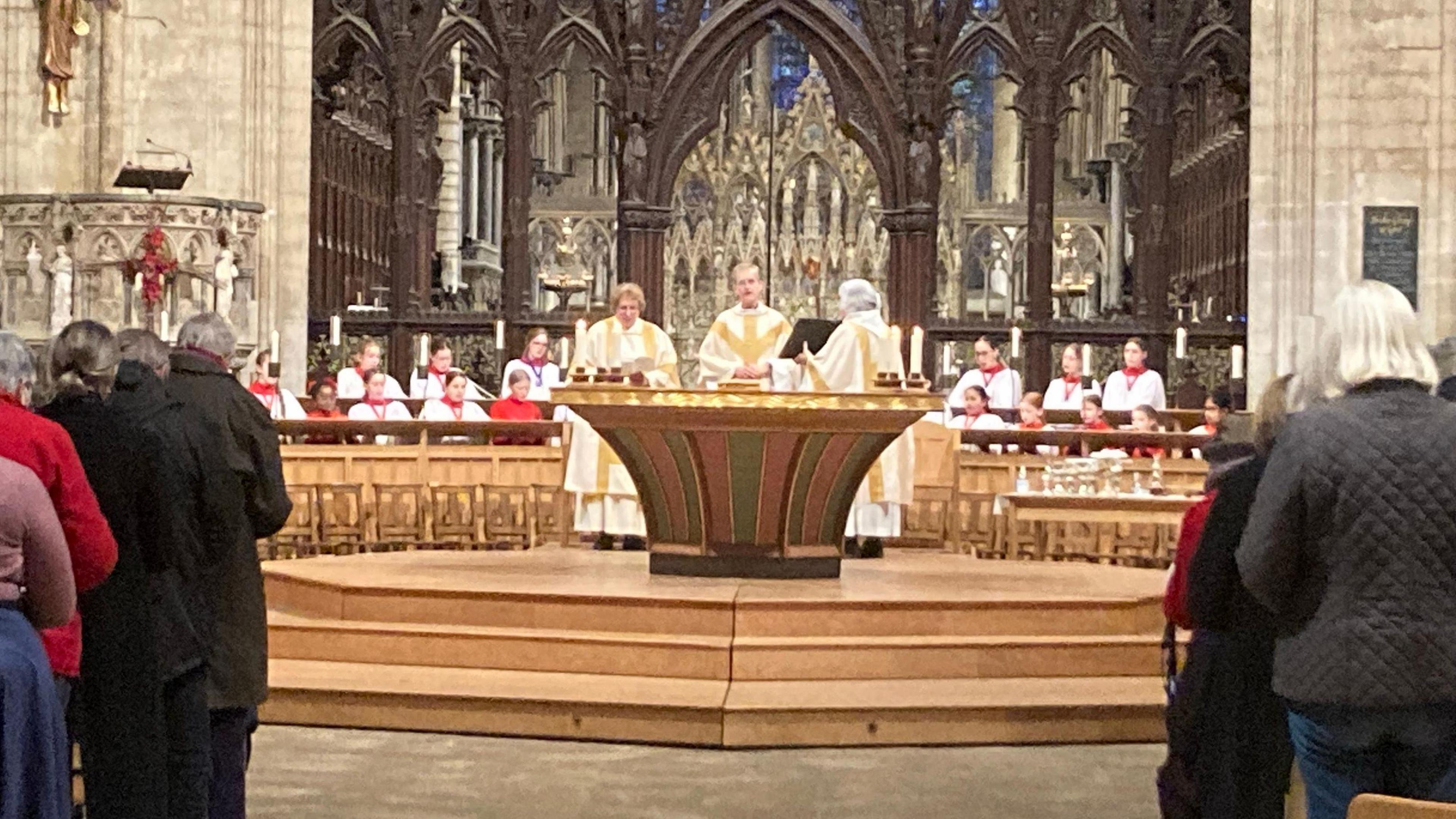 A church service at Ely Cathedral. Members of the congregation standing either side of the aisle, seen from behind. The alter at the front has 3 priests standing behind and facing the congregation, two women and a man. They are wearing white robes with gold detail. A choir sits behind them wearing white robes and red detail. In the background the stone building, with a pulpit on the left and brown wooden arches in the centre behind the choir.