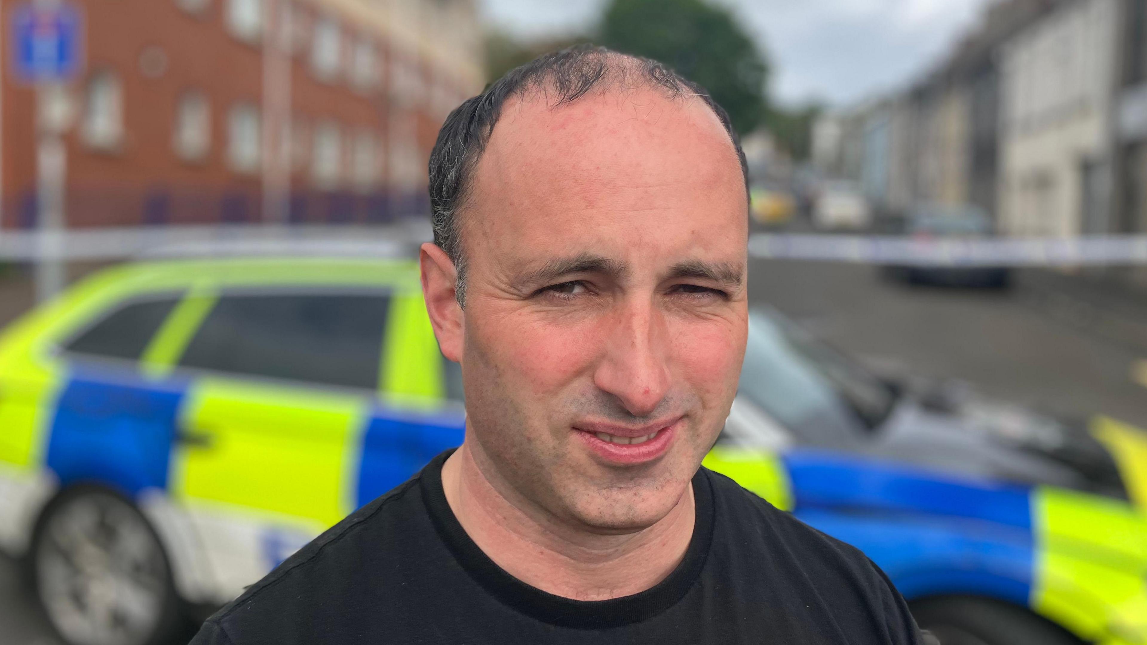 DUP Councillor Andrew Clarke standing in front of a police car. He has dark, thinning hair and is wearing a black t-shirt