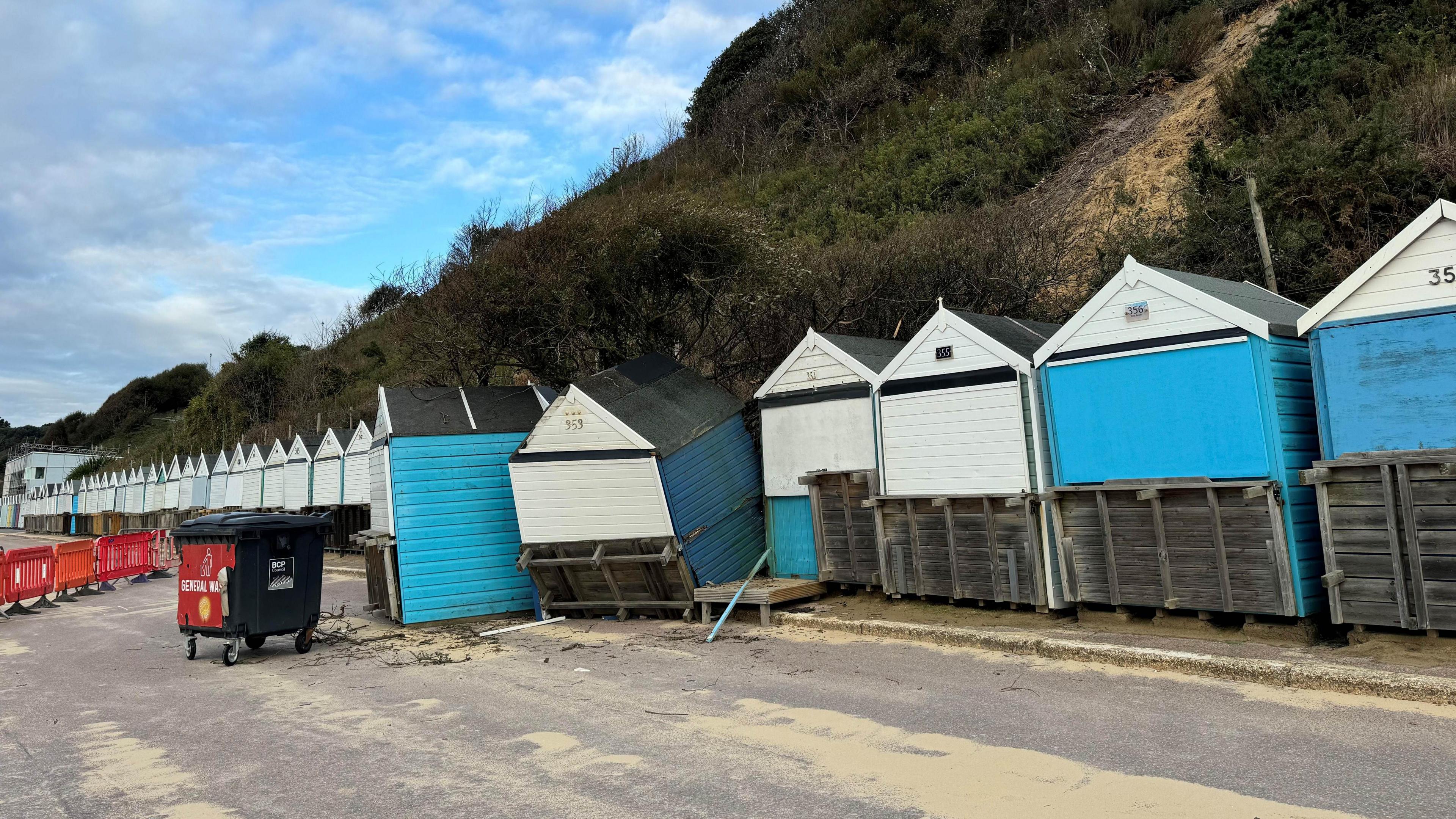 Beach huts at Durley Chine, which are wooden with pointed roofs. Behind them is a cliff. Two of the beach huts have been pushed out of line