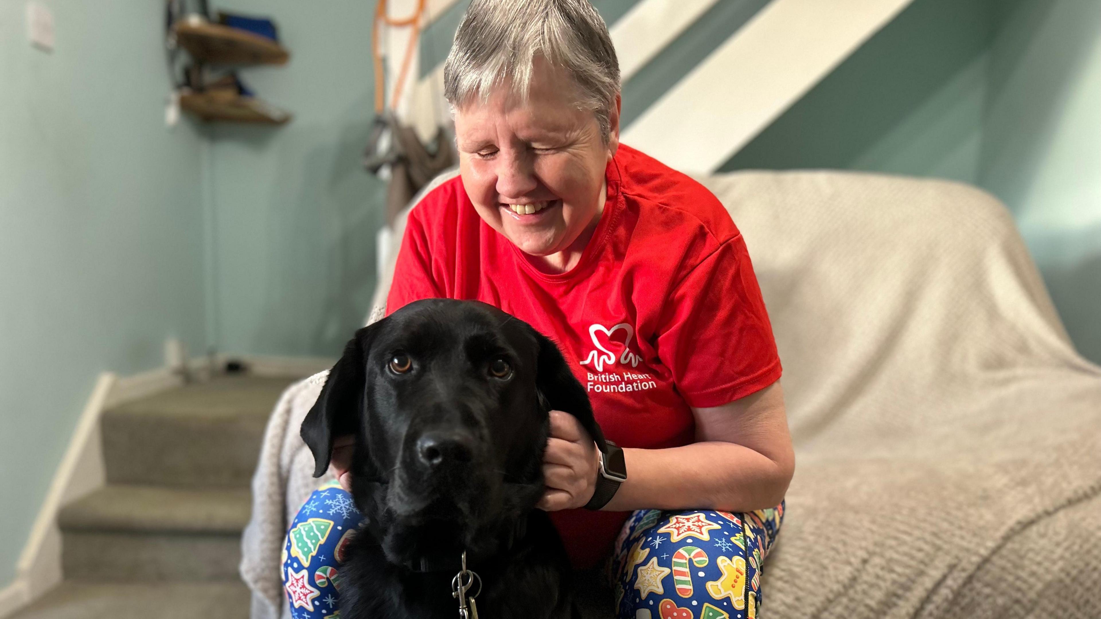 A blind woman in a red British Heart Foundation t-shirt pictured with her black Labrador guide dog, which is looking straight at the camera .She is sitting on an armchair which has a beige blanket draped across it and also wearing blue leggings with a colourful print of candy canes, gingerbread men and stars. A staircase and bannister are out of focus behind her and the walls are painted blue.