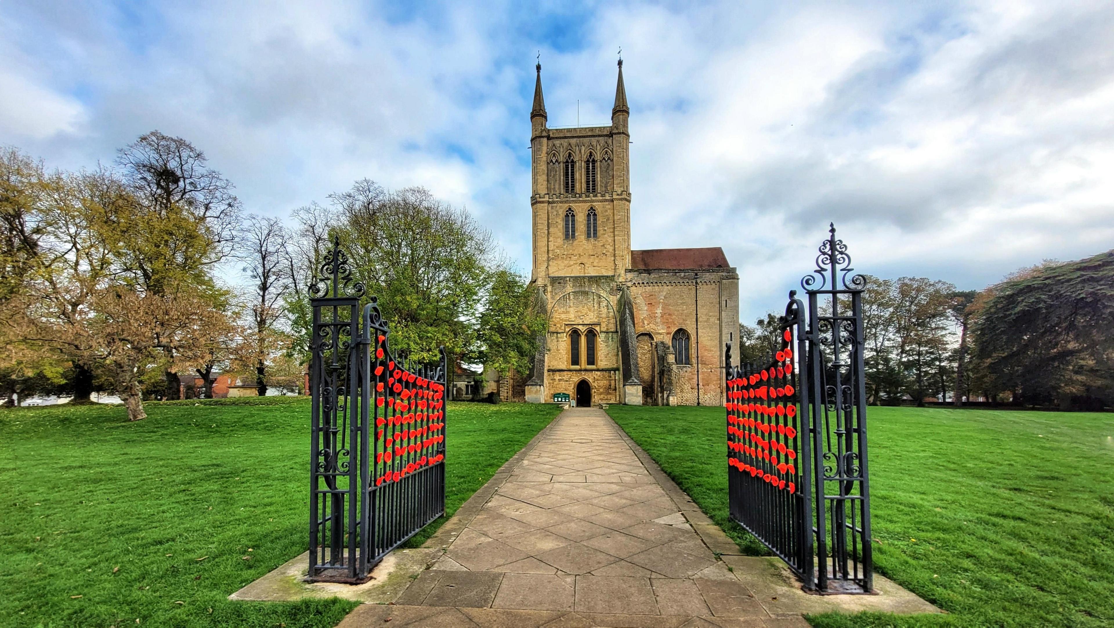 Wrought-iron gates at the entrance to a former Benedictine abbey surrounded by grass. The gates have been adorned with dozens of red Remembrance poppies. 