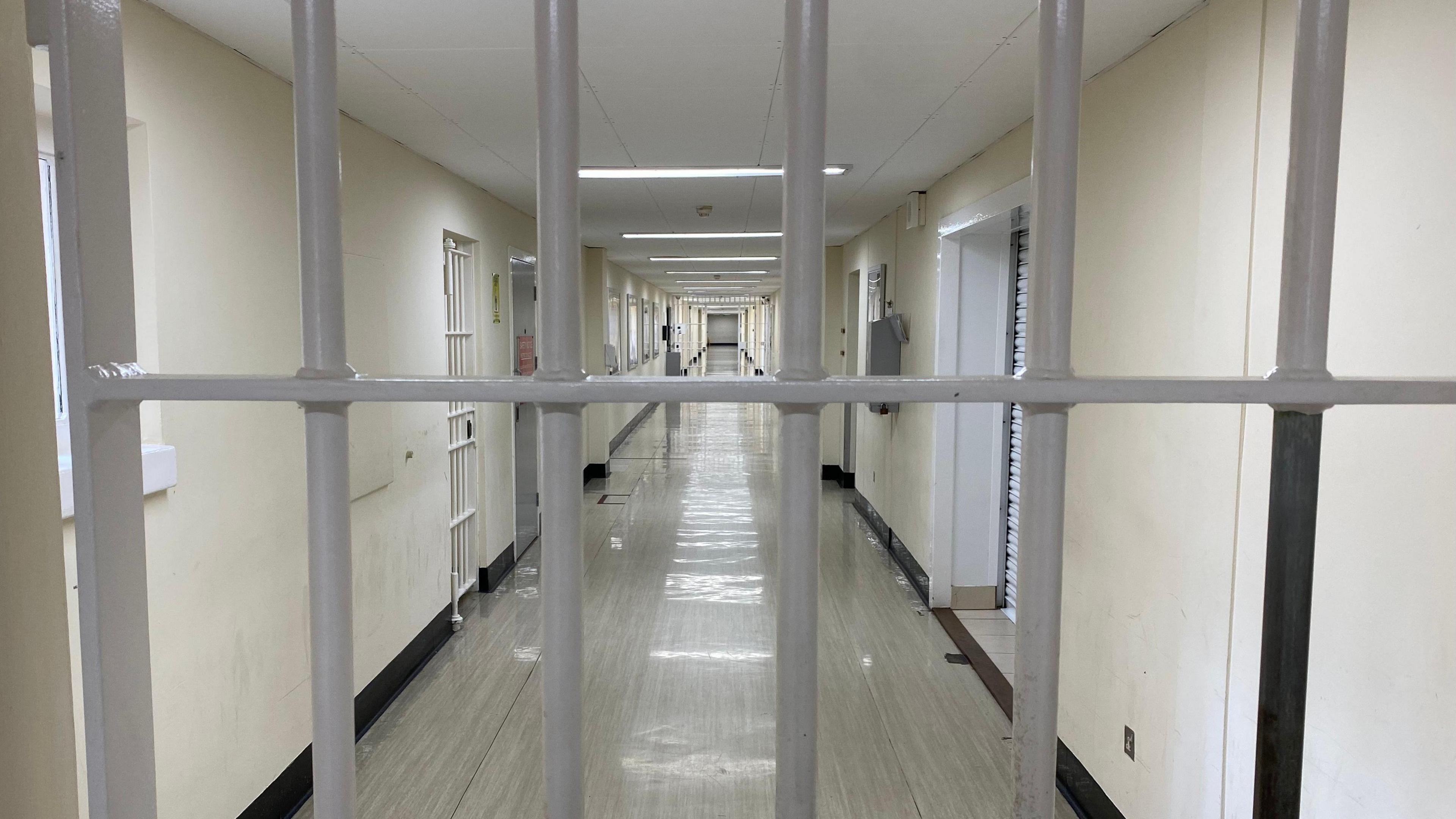 The camera looks down a long prison corridor, with a grey lino floor and what appear to be doors, with bars across them. There is a grey barred door in the front of the image, blocking the photographer's access.