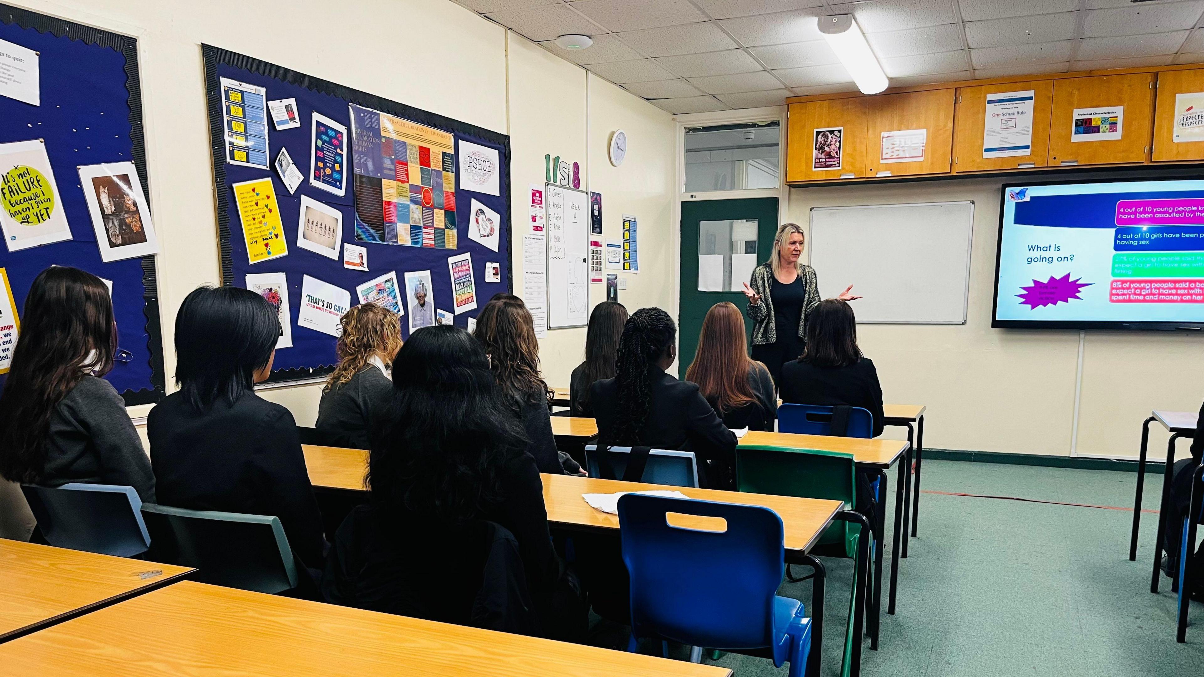 Mandy Geraghty stands in front of a class in a classroom and is talking to students who are sitting at rows of tables wearing a dark-coloured uniform. She is standing next to a smartboard with a power-point presentation on it.
