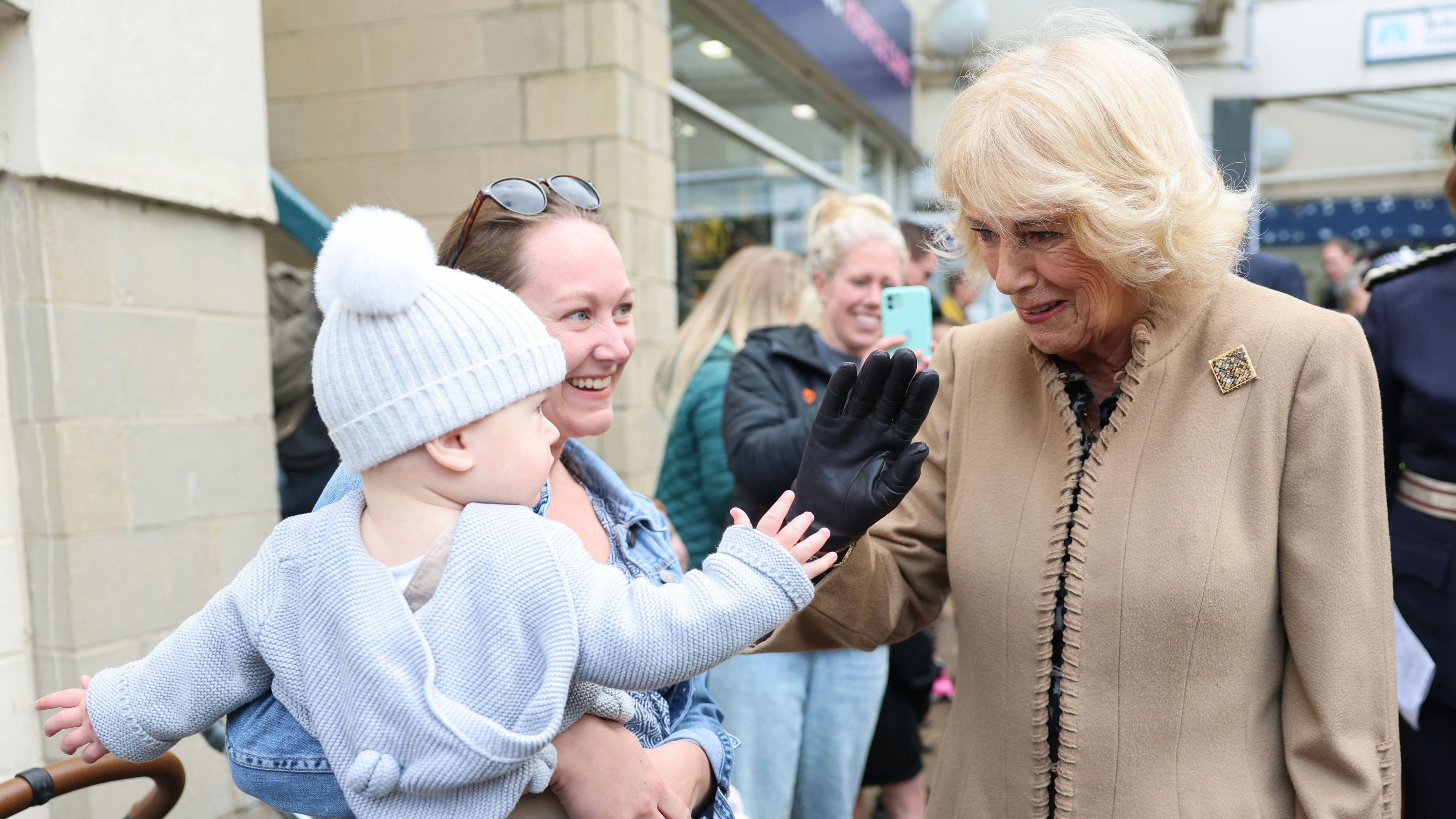 Queen Camilla raises a black gloved hand to high five a small baby in a bobble hat being held by a woman with brown hair